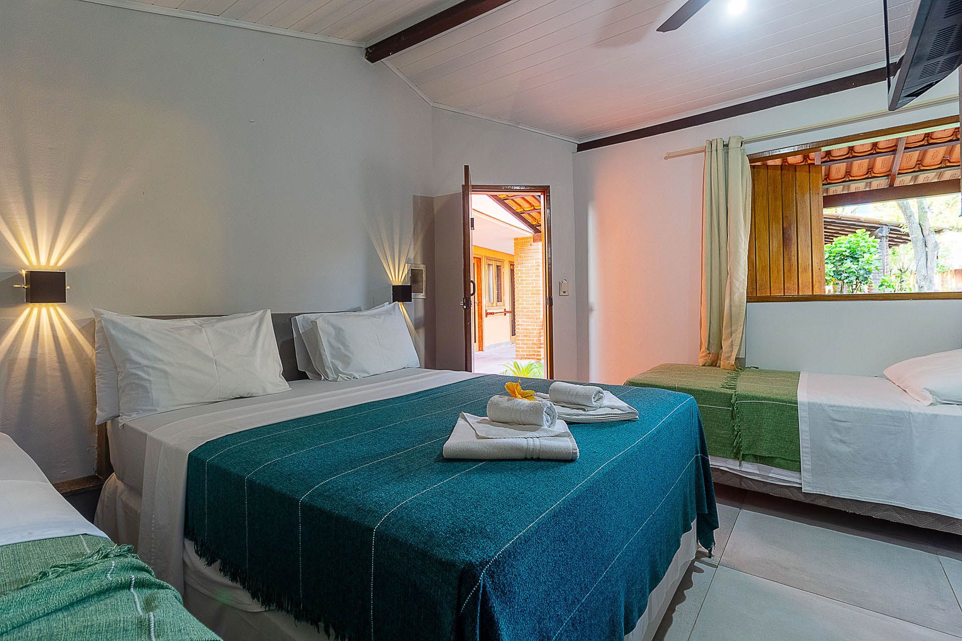 Quarto de hotel luminoso com duas camas, colcha azul-petróleo, apliques de parede, ventilador de teto e janela aberta.