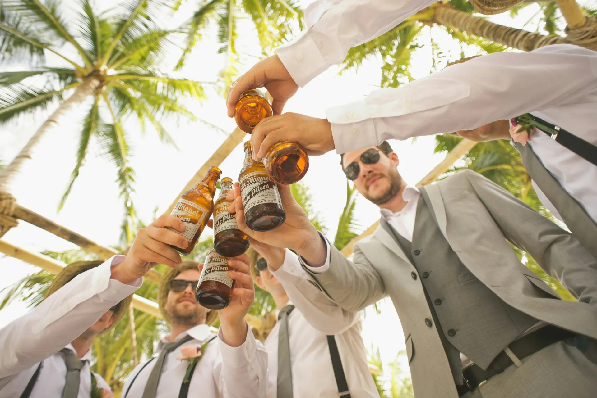 Groomsmen in suits cheer with beer bottles under palm trees.