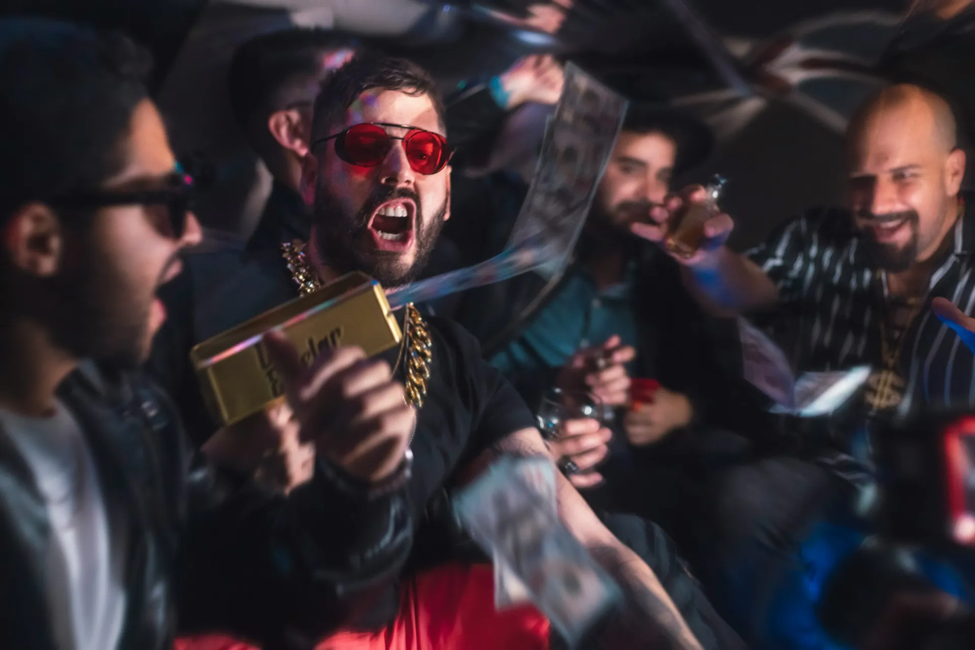 Man in red sunglasses holds a gold box shooting money and yells in crowded setting.