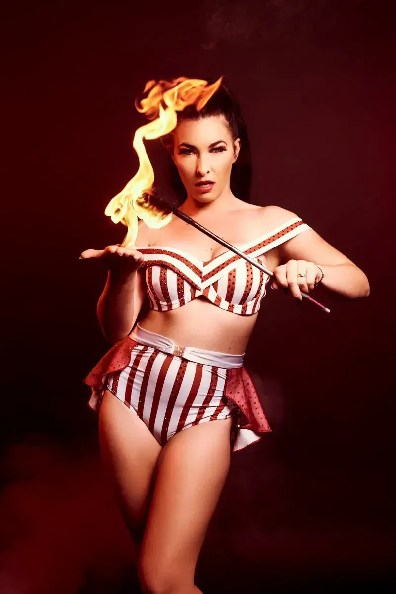 Fire dancer in striped costume holding wand and flame against a red background.