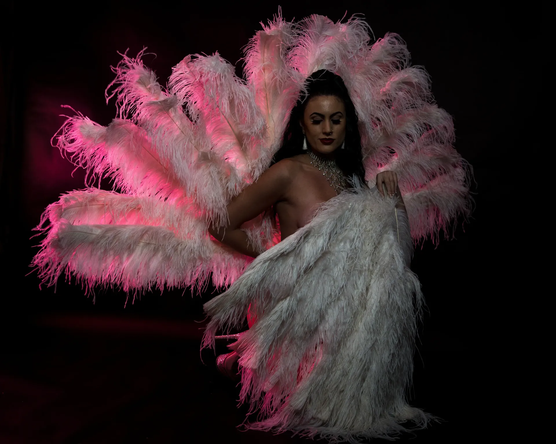 Woman in a white feathery costume against a dark background, illuminated by pink light.