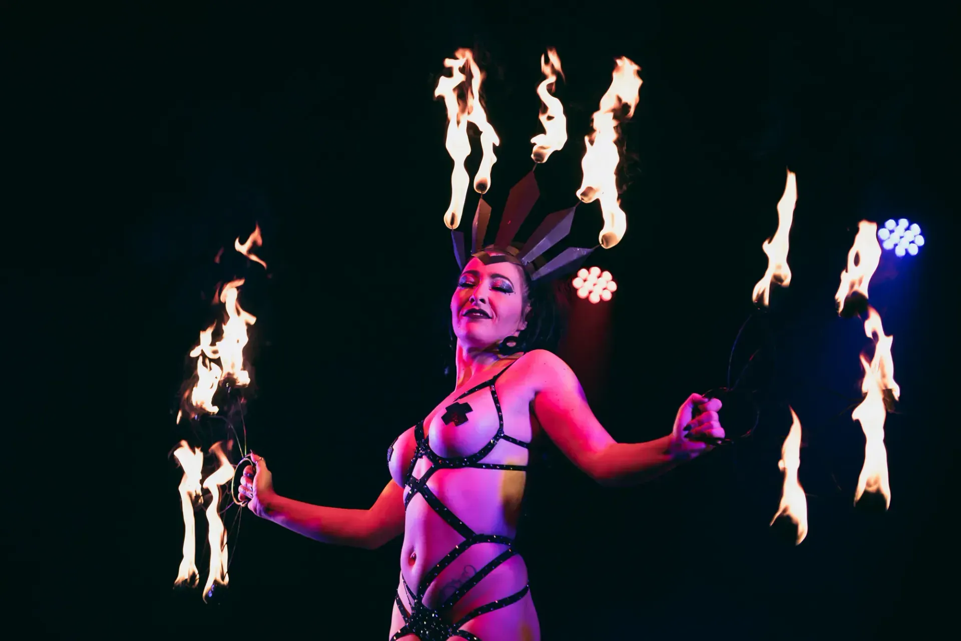 Fire dancer in a flame crown headpiece, posing on stage while holding fire props with flames burning beside her.