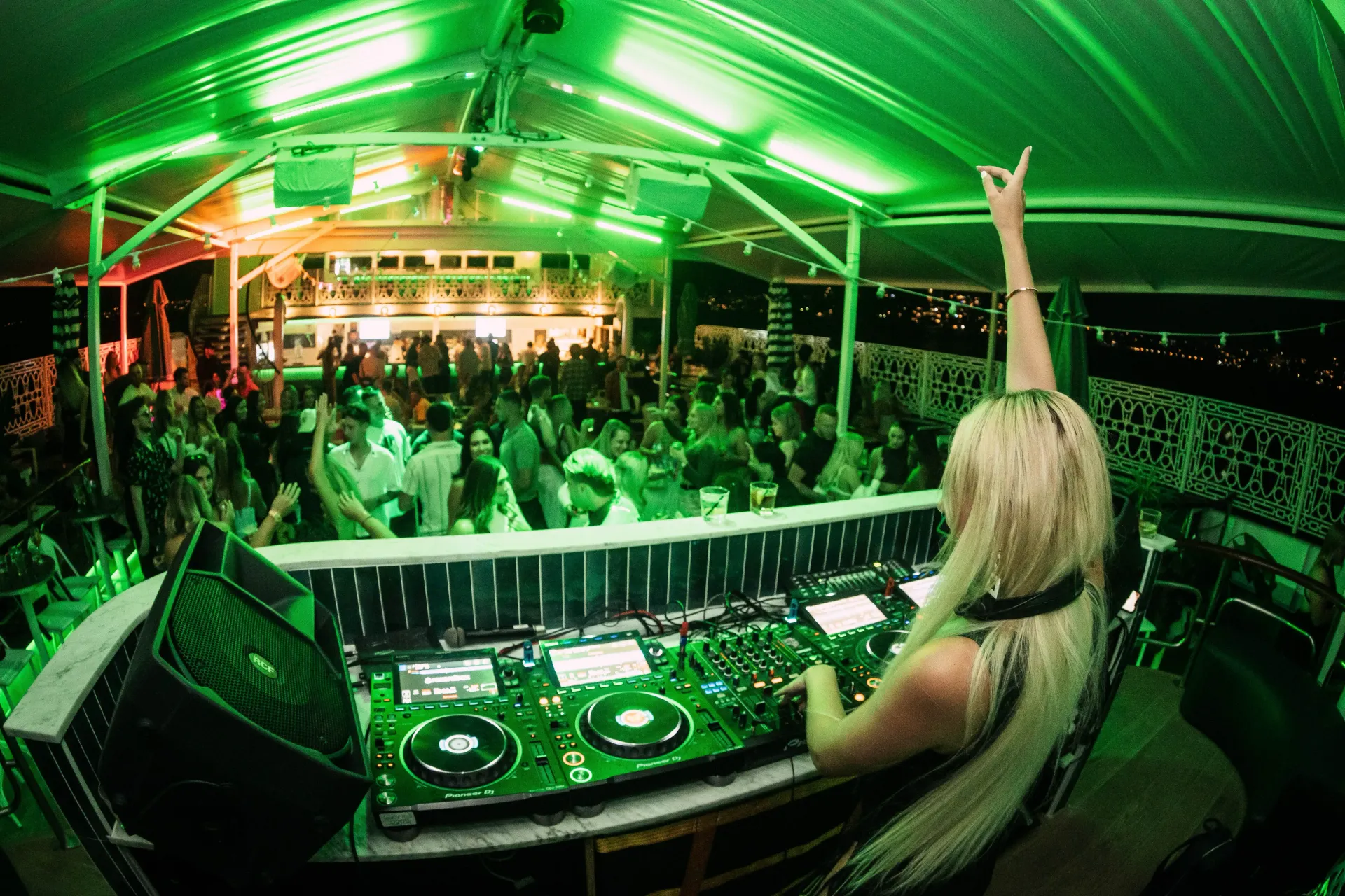 Blonde DJ at a green-lit outdoor club, arms raised, mixing music for a crowd.