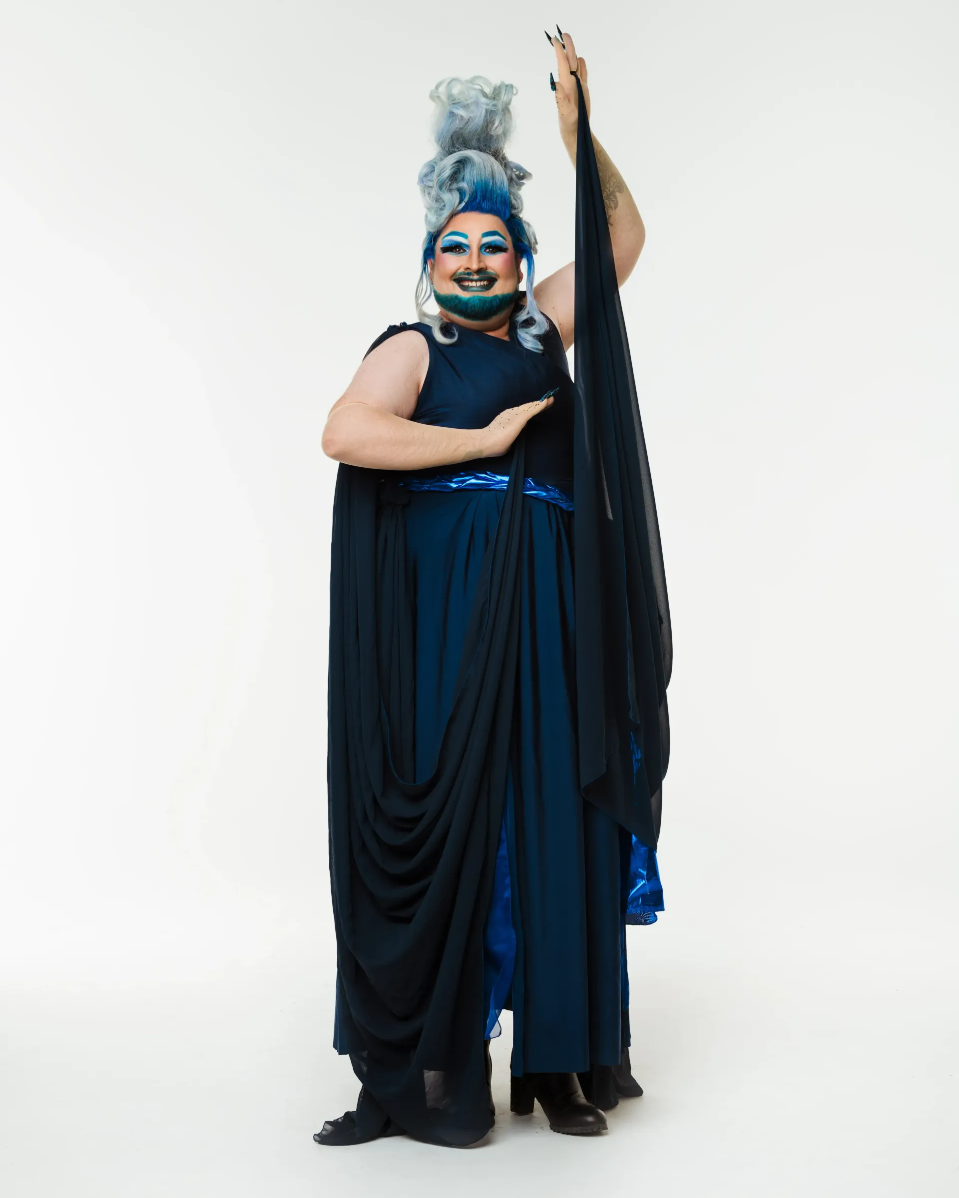 Drag performer in dark blue dress, posing arm up, vibrant make up on white background.