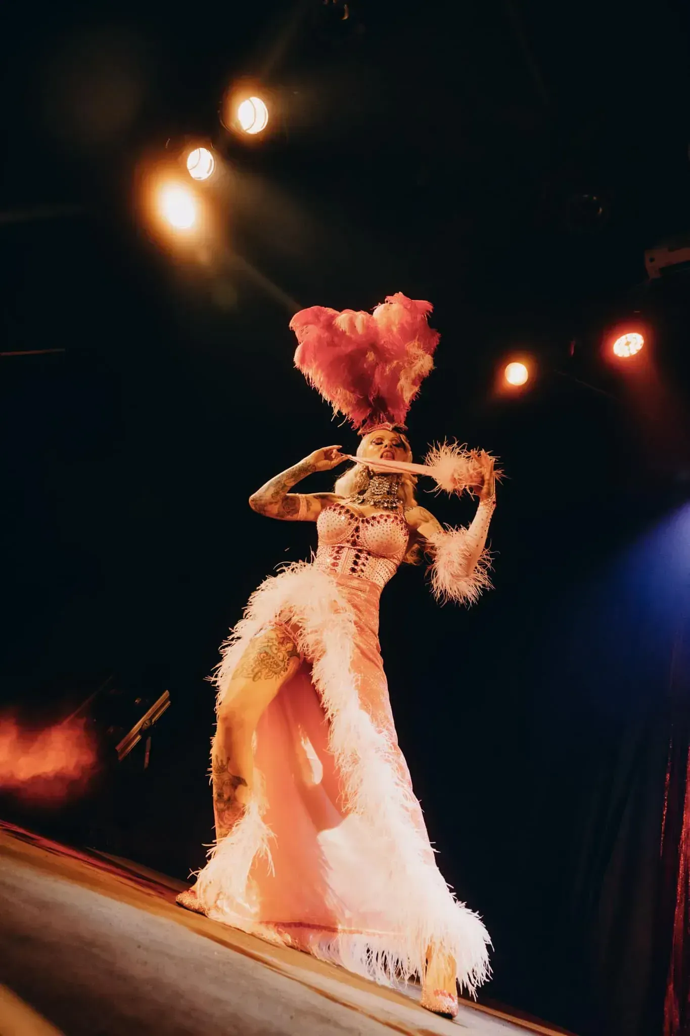 Burlesque performer in feathery pink gown and heart-shaped headdress on stage.
