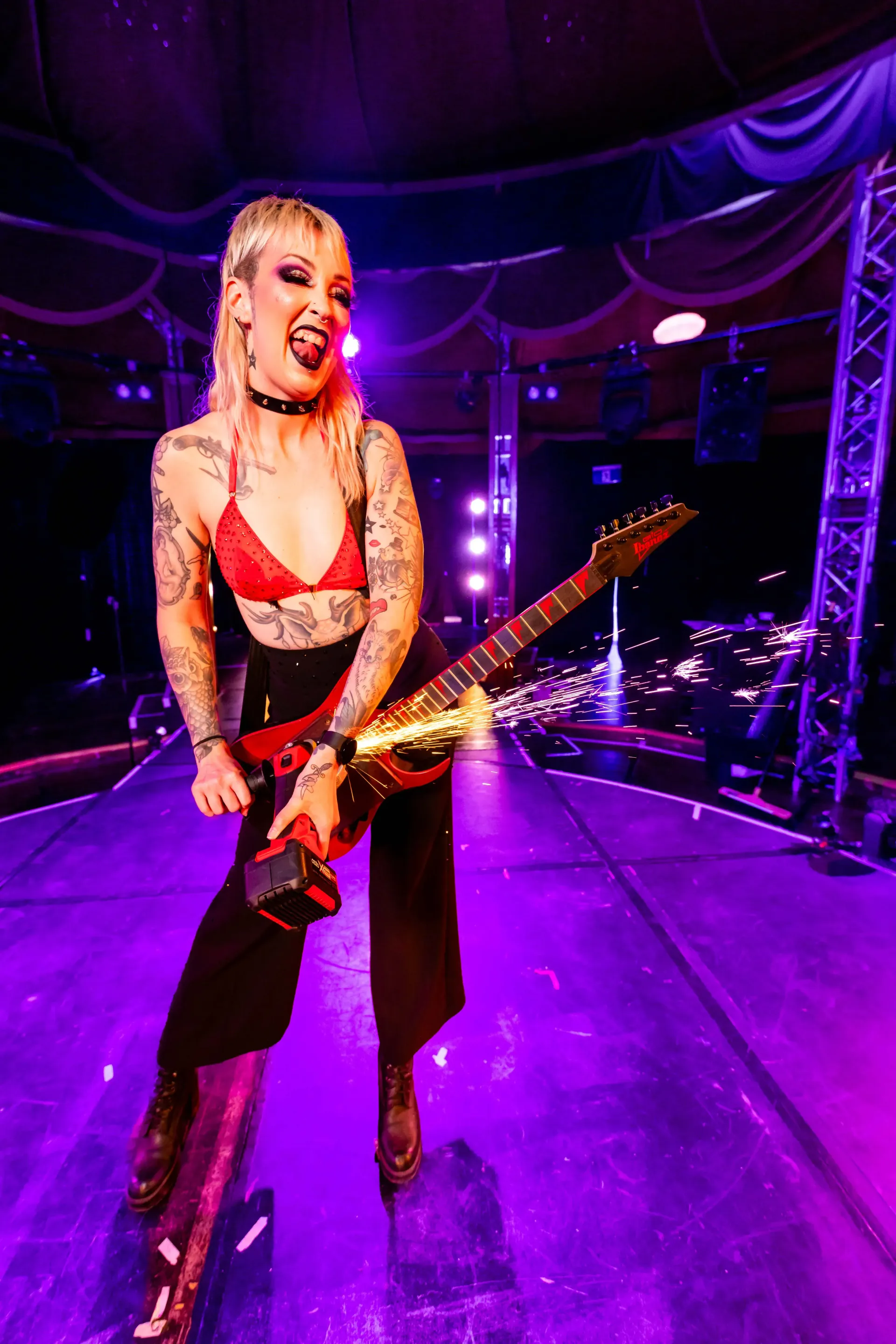 Fire dancer with tattoos, red top, and black pants, playing a red and gold guitar on a stage.