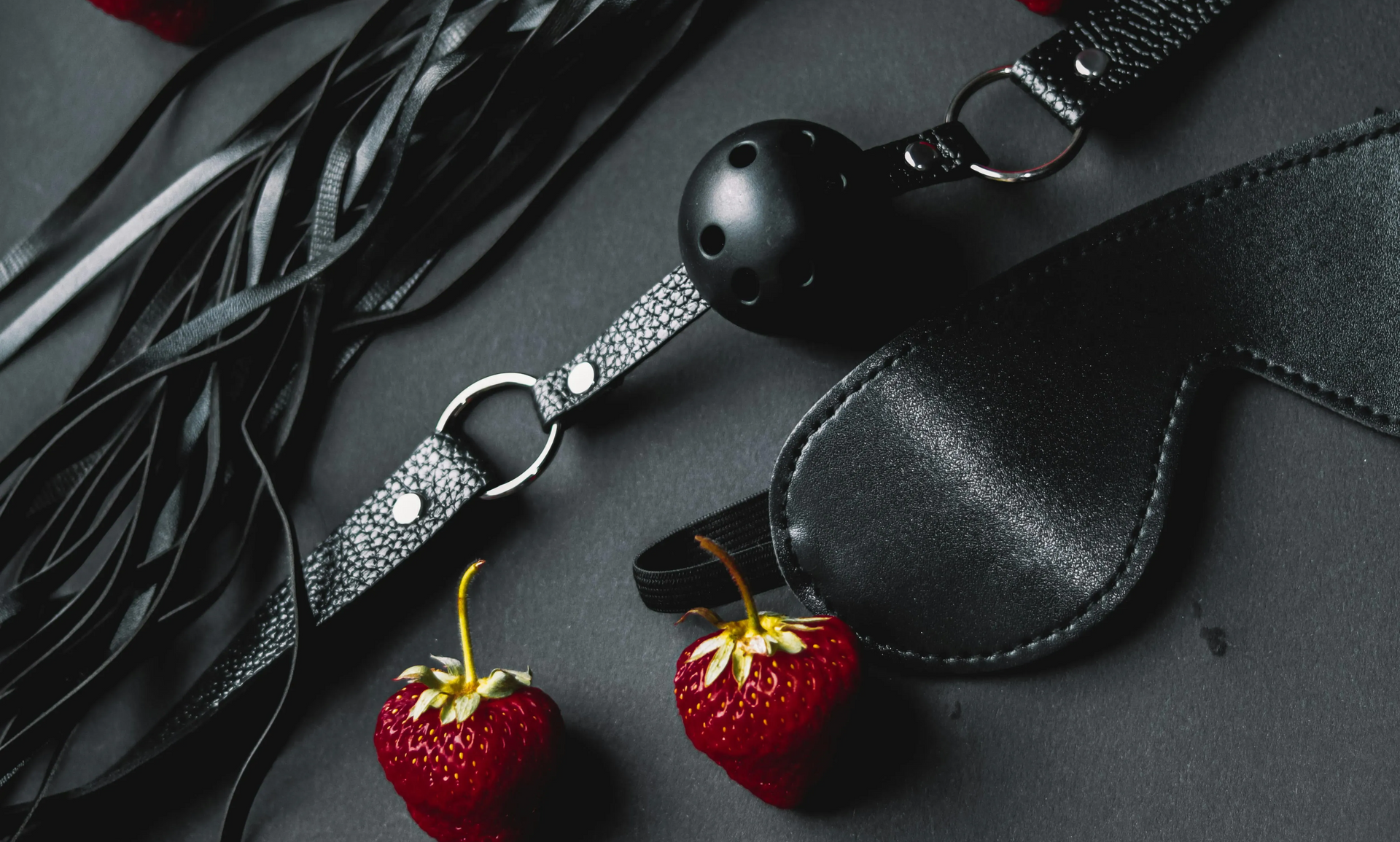 A ball gag, a leather whip, and an eye mask arranged on a dark surface with two fresh strawberries.