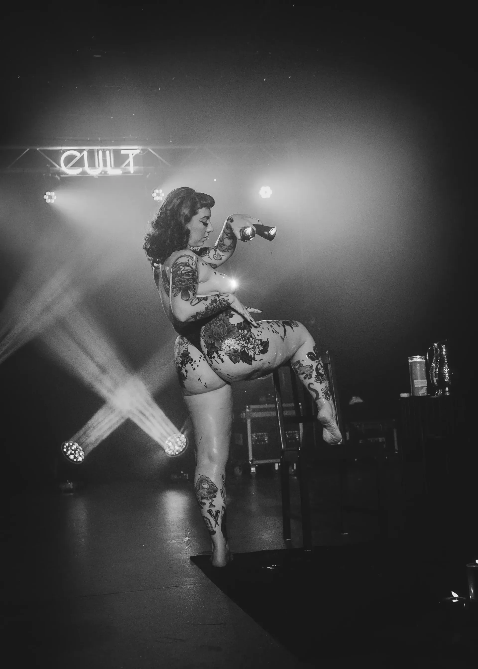 Burlesque performer on stage, covered in tattoos and wax kicking leg up.