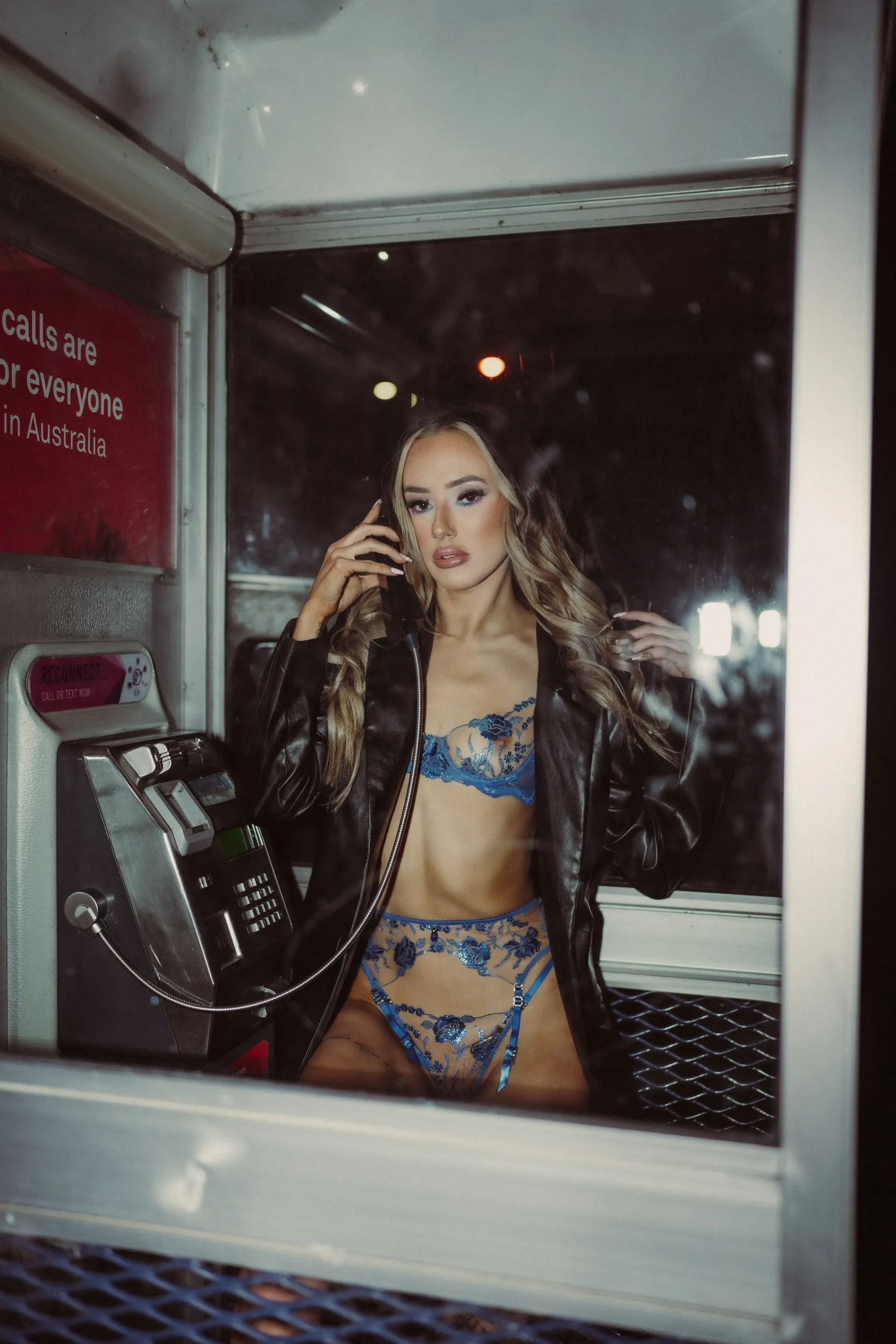 Topless waitress in blue lace lingerie and a black leather jacket poses inside a telephone booth.