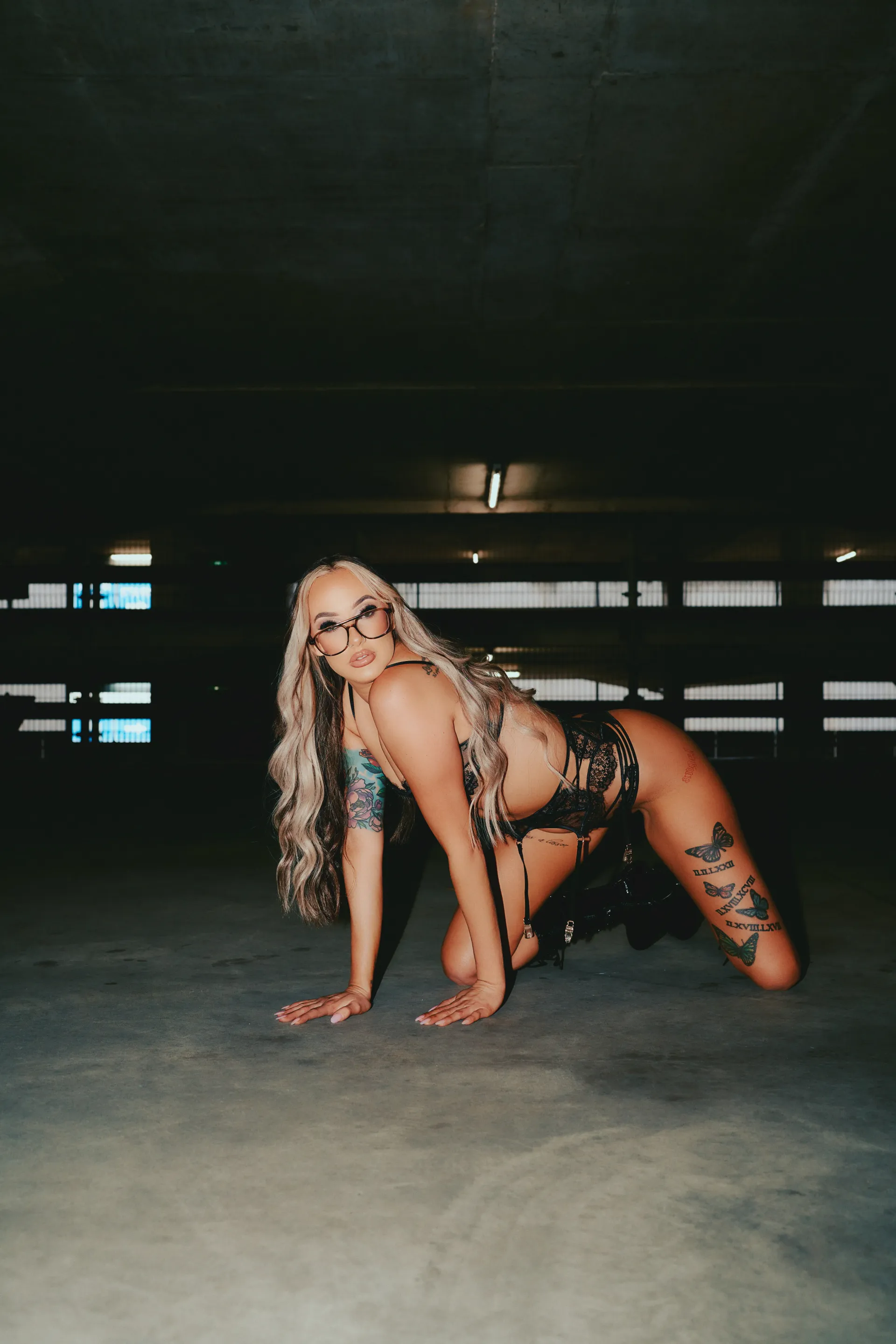 A lingerie waitress with long, blonde, wavy hair in a dark, patterned two-piece outfit posing on the concrete floor of a garage.