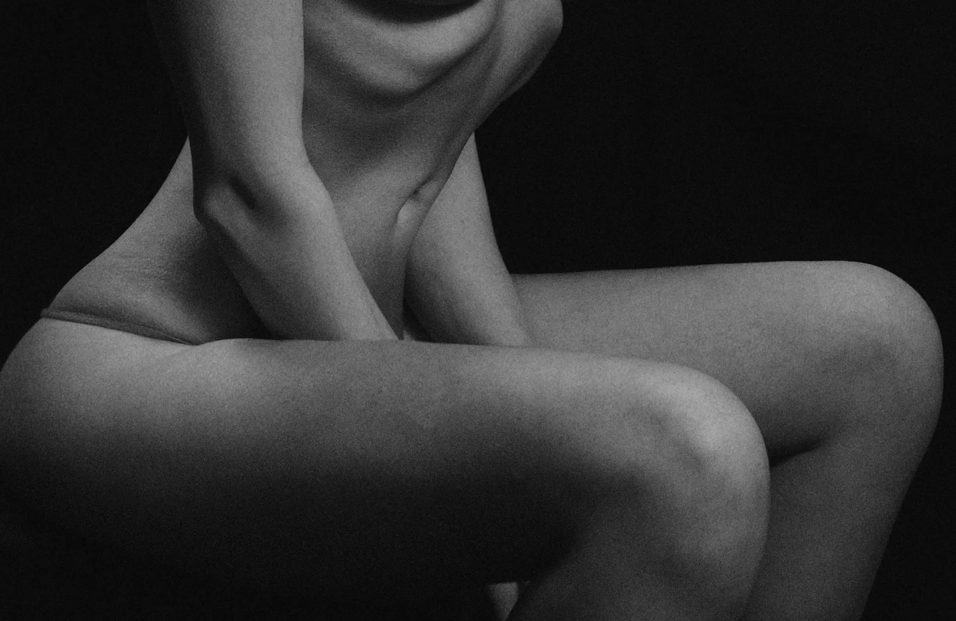 Black-and-white close-up of a seated nude person, cropped at the torso and thighs against a dark background