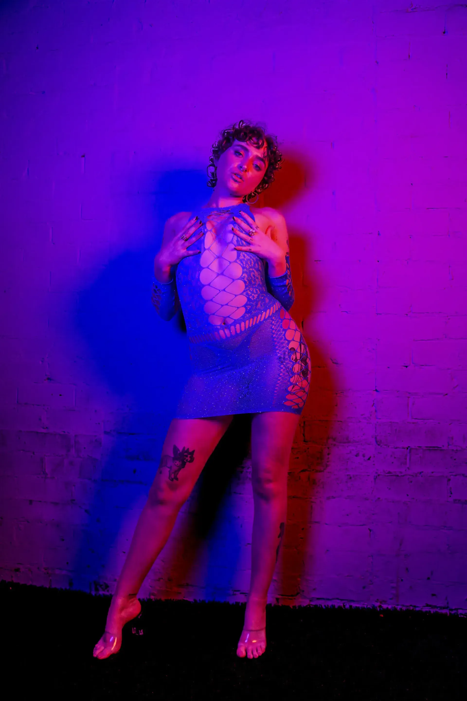 Stripper in blue dress, posing against purple wall, arms up, legs crossed, wearing clear heels.