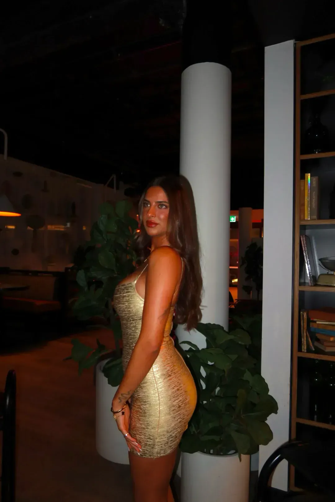 Woman in a gold dress posing at night beside a white column and potted plant.