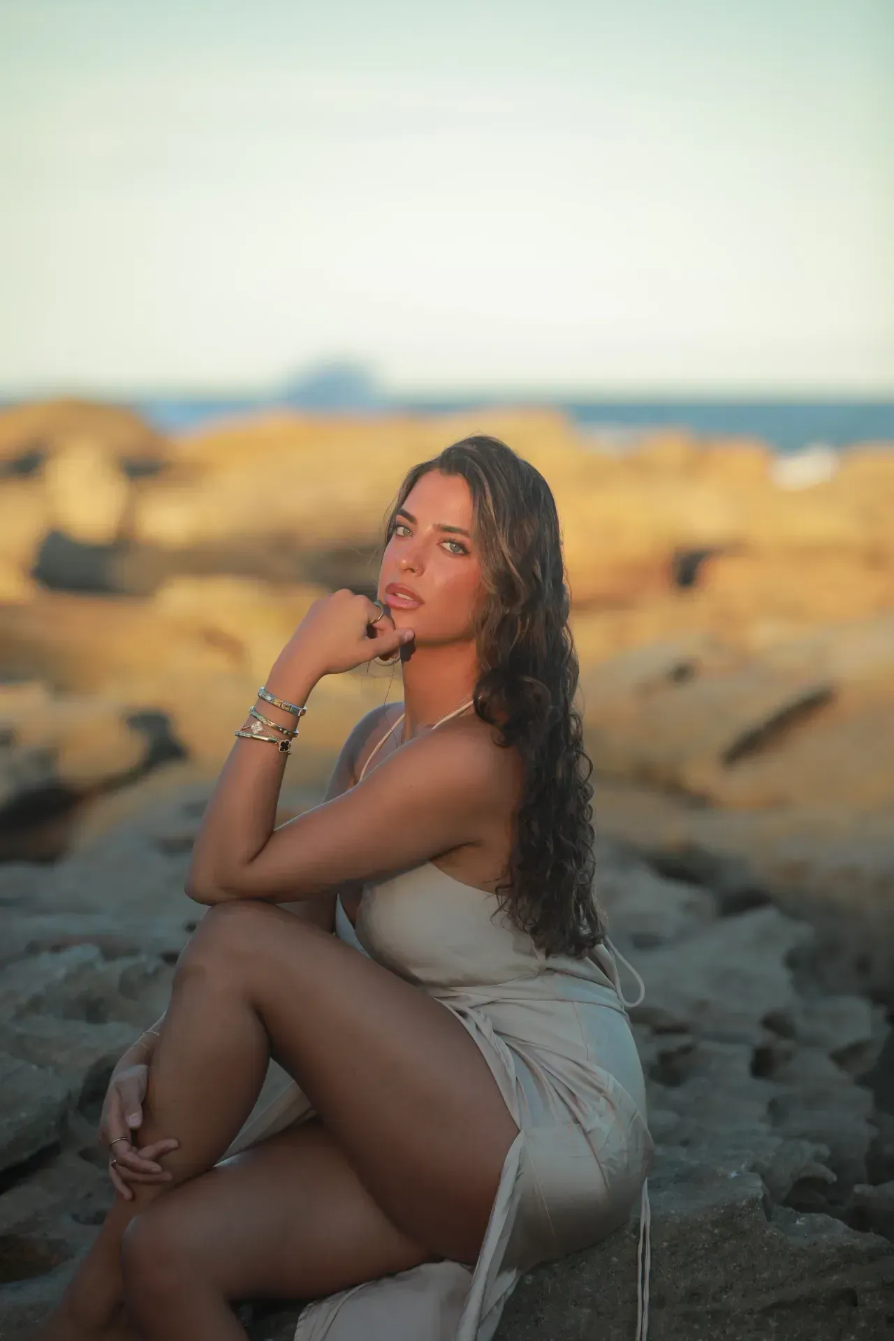 Woman seated on rocky shore at sunset, wearing a white top and gray skirt, looking out over the water