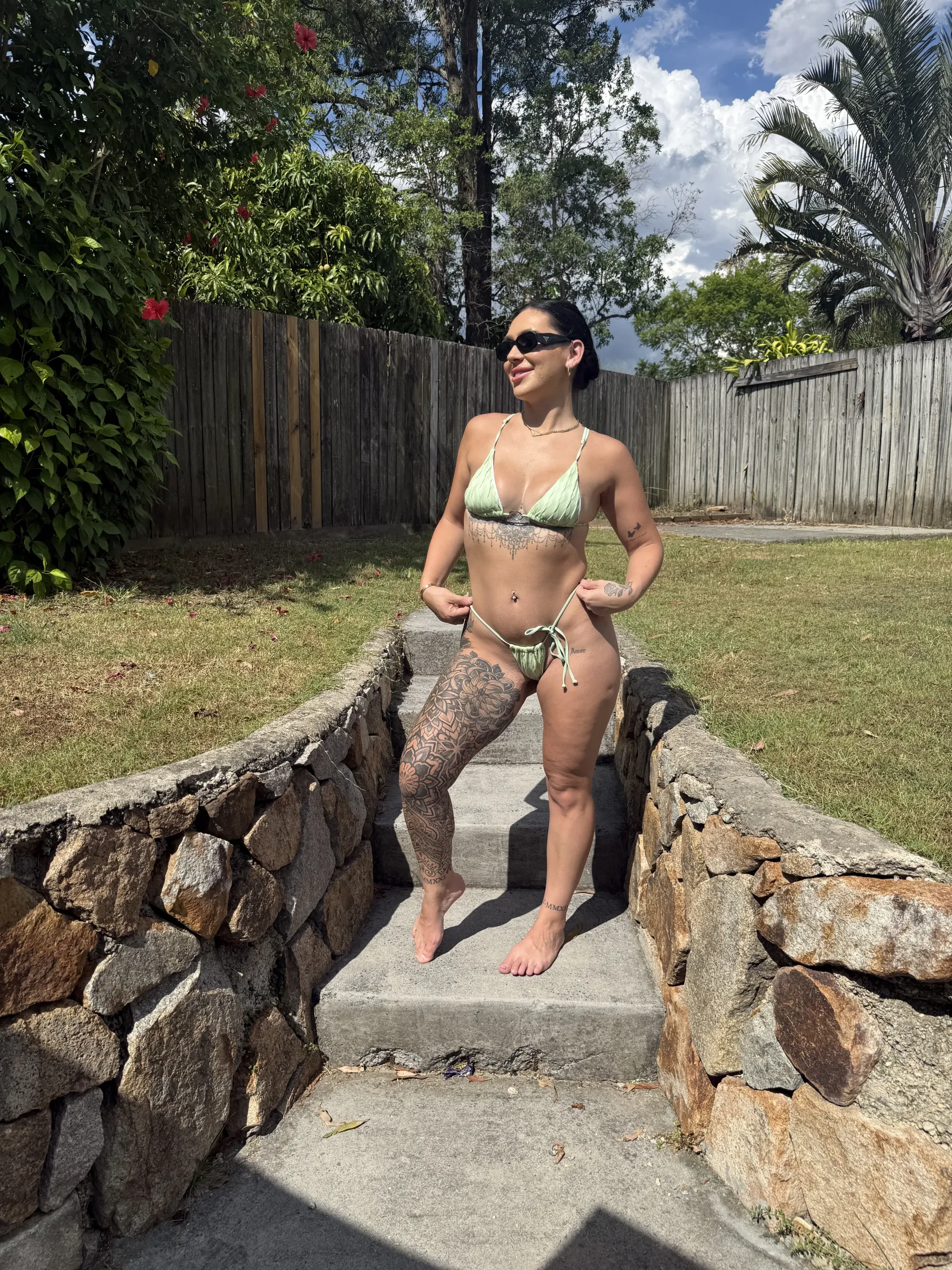Bikini waitress wearing a green bikini and sunglasses stands on stone steps, smiling in a sunny outdoor setting.