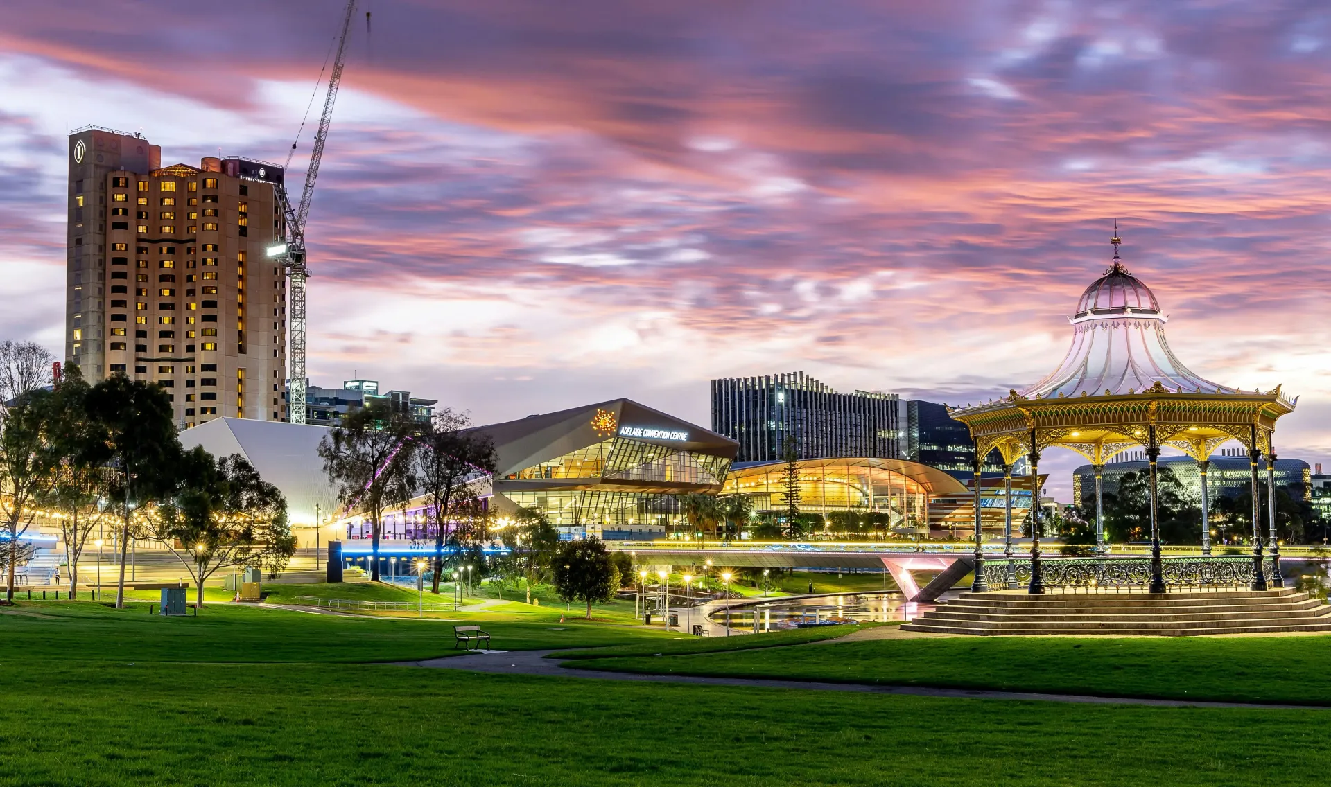 City park at dusk with lit buildings, a domed pavilion, and pink-purple clouds over green grass