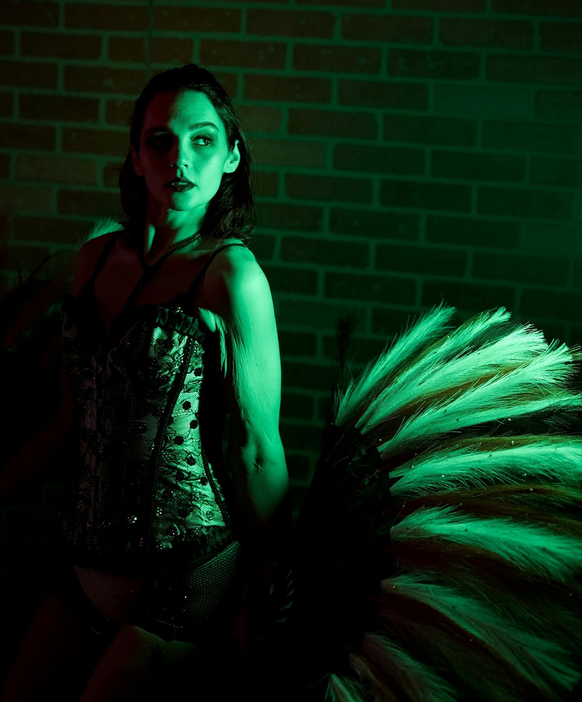 Moody green-lit burlesque performer in a corset holding large feather fan, posed against a brick wall backdrop.