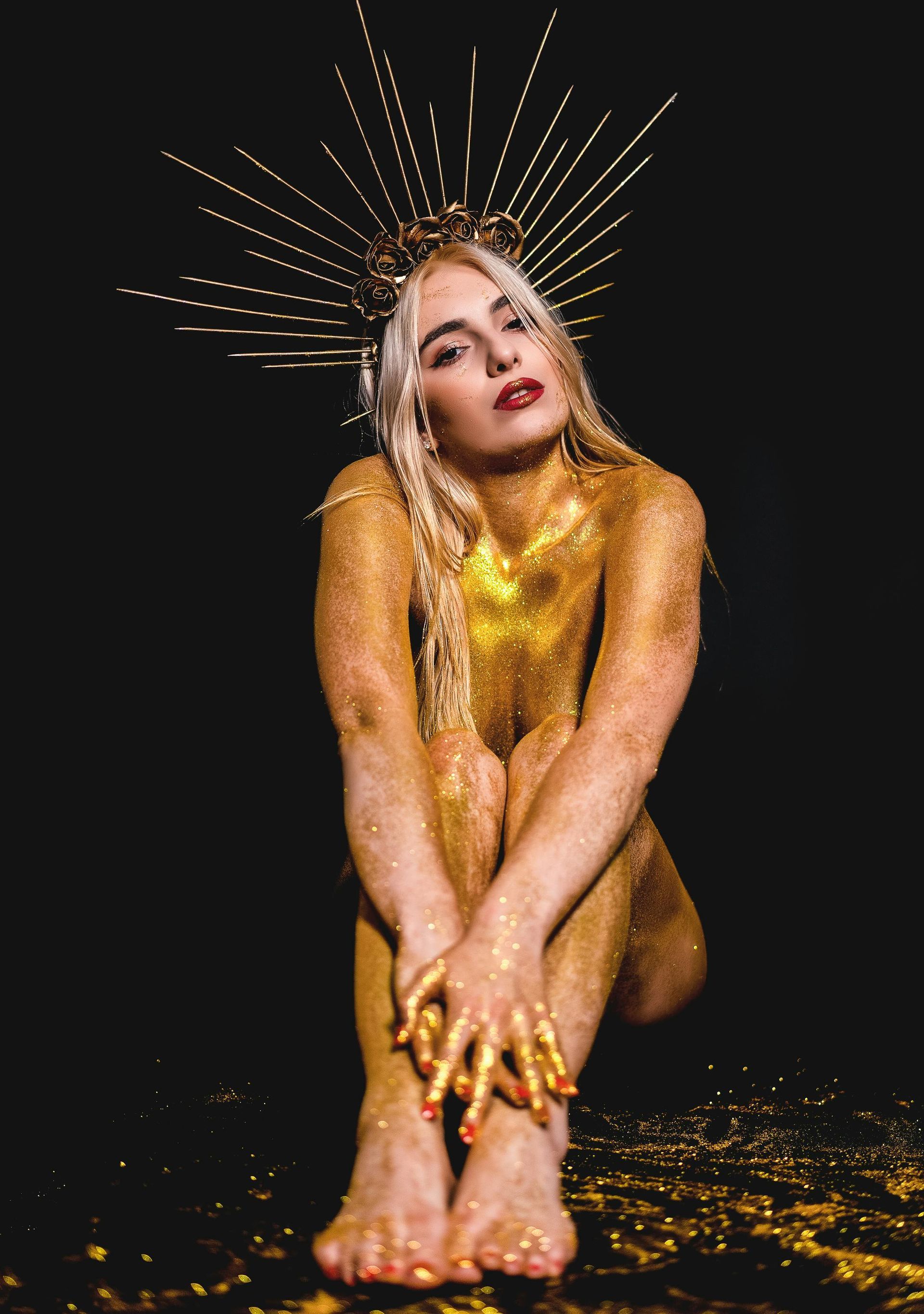 Gold-glitter burlesque performer seated on a dark set, wearing a radiant spiked halo headpiece and bold red lipstick.