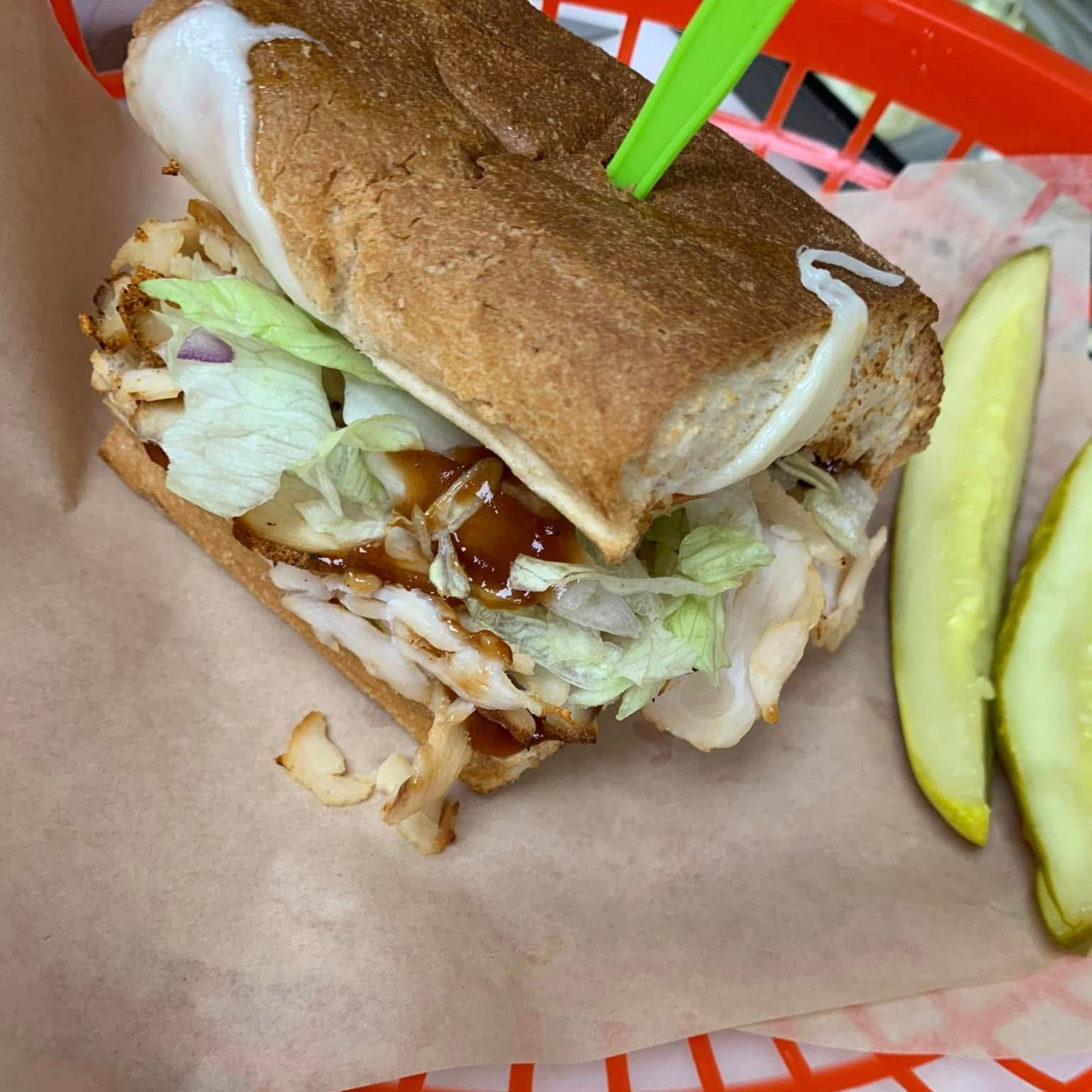 Sub sandwich with sliced meat, lettuce, and sauce, served with pickles in a red basket.