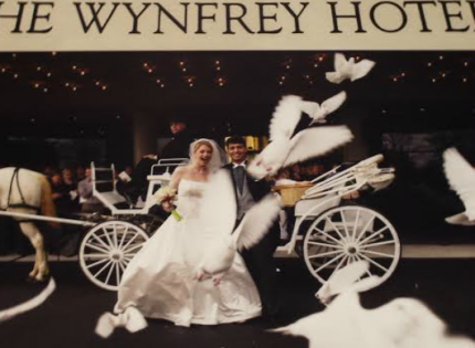 Bride in white gown standing by a white horse-drawn carriage at The Wynfrey Hotel, with white birds flying around