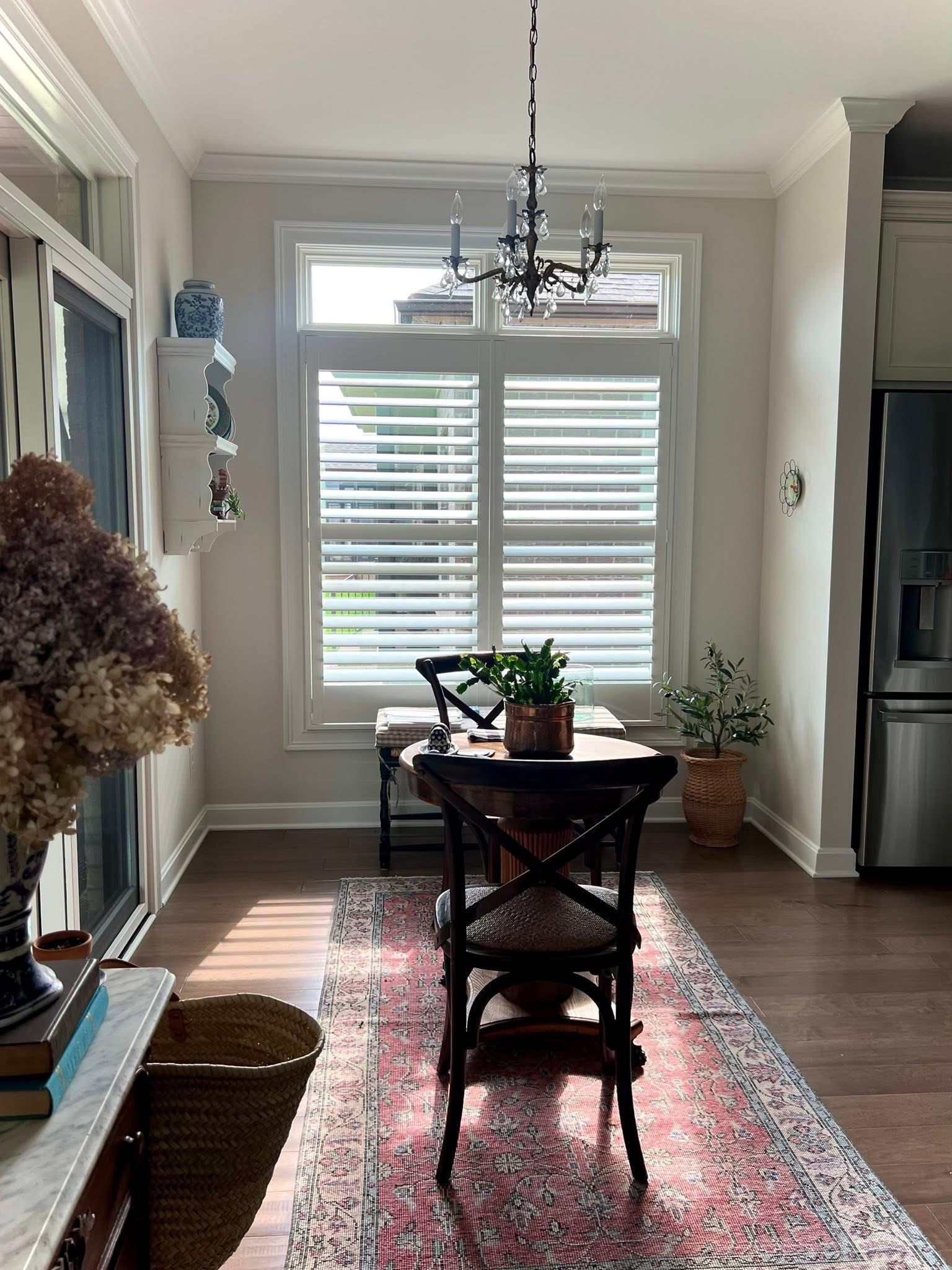 Dining room with table, chairs, rug, and shuttered window; sunlight streaming in.