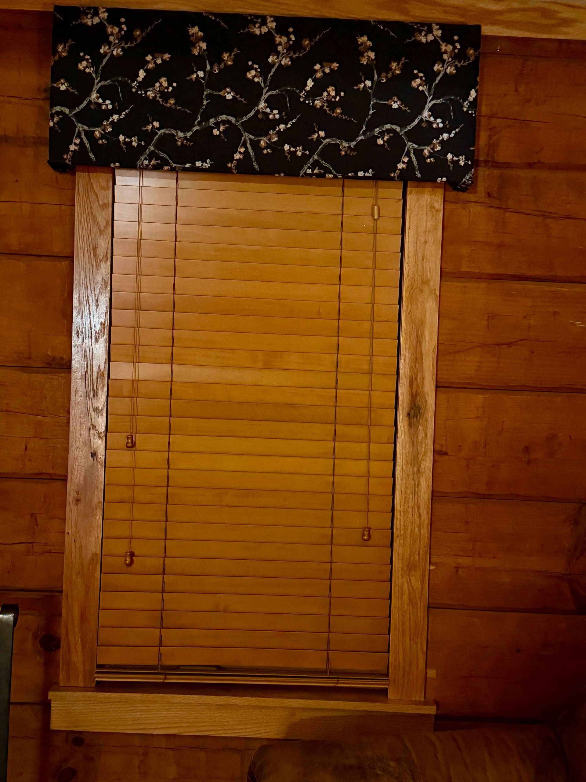 Wooden window with blinds, topped by a black floral valance, set in a wood-paneled wall.