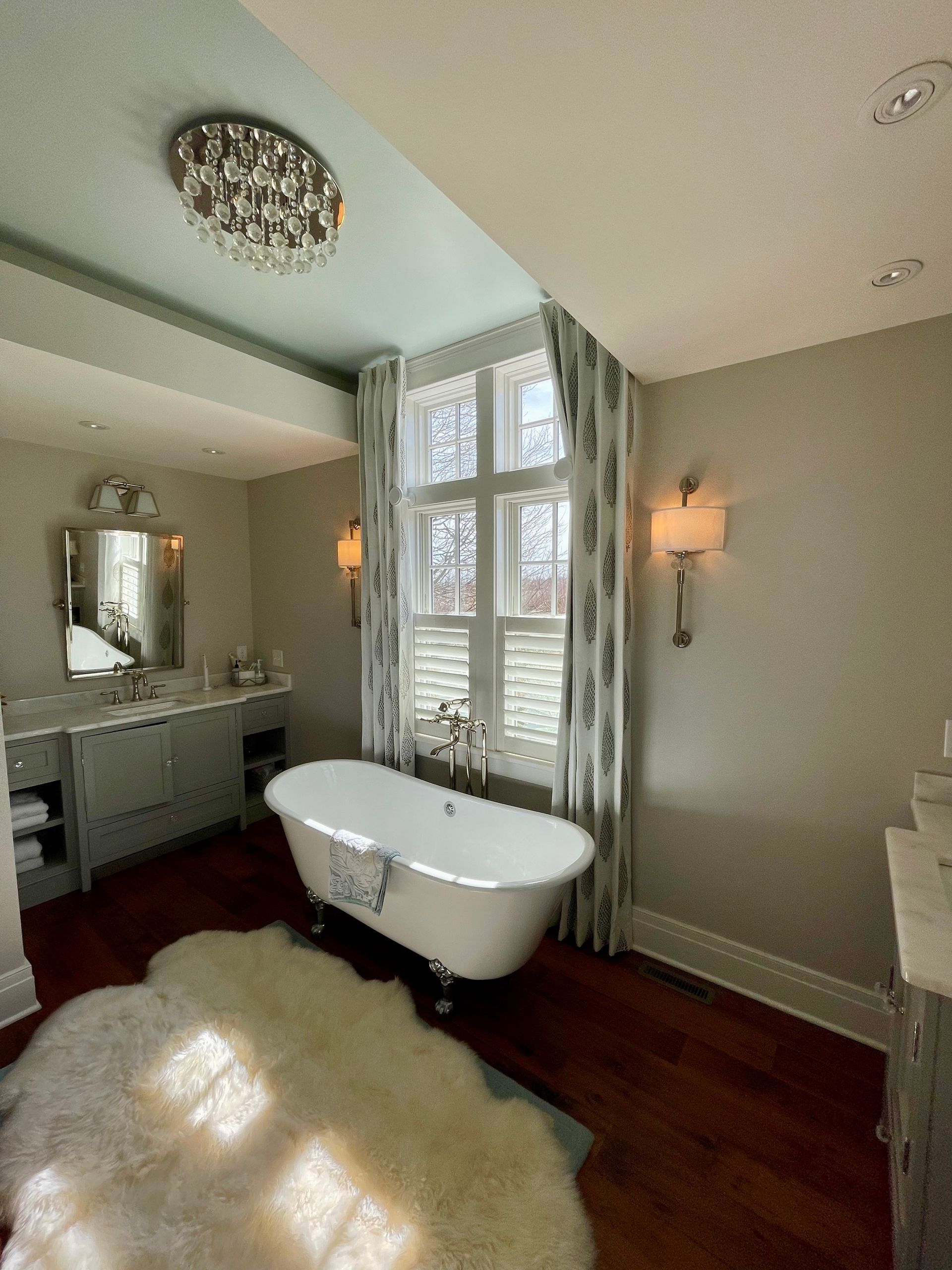 Bathroom with a clawfoot tub, rug, and vanity; light blue ceiling, white walls, and a window with curtains.