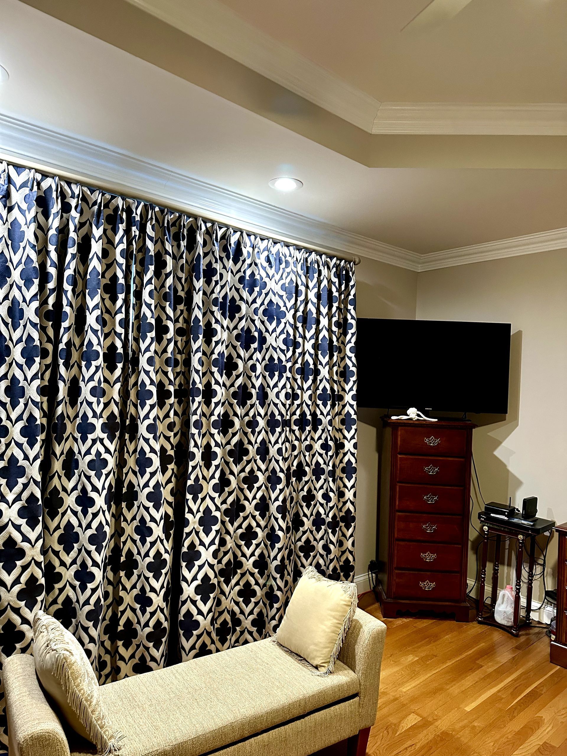 Navy and white patterned curtains frame a bench seat, with a TV, chest of drawers, and crown molding in a room.