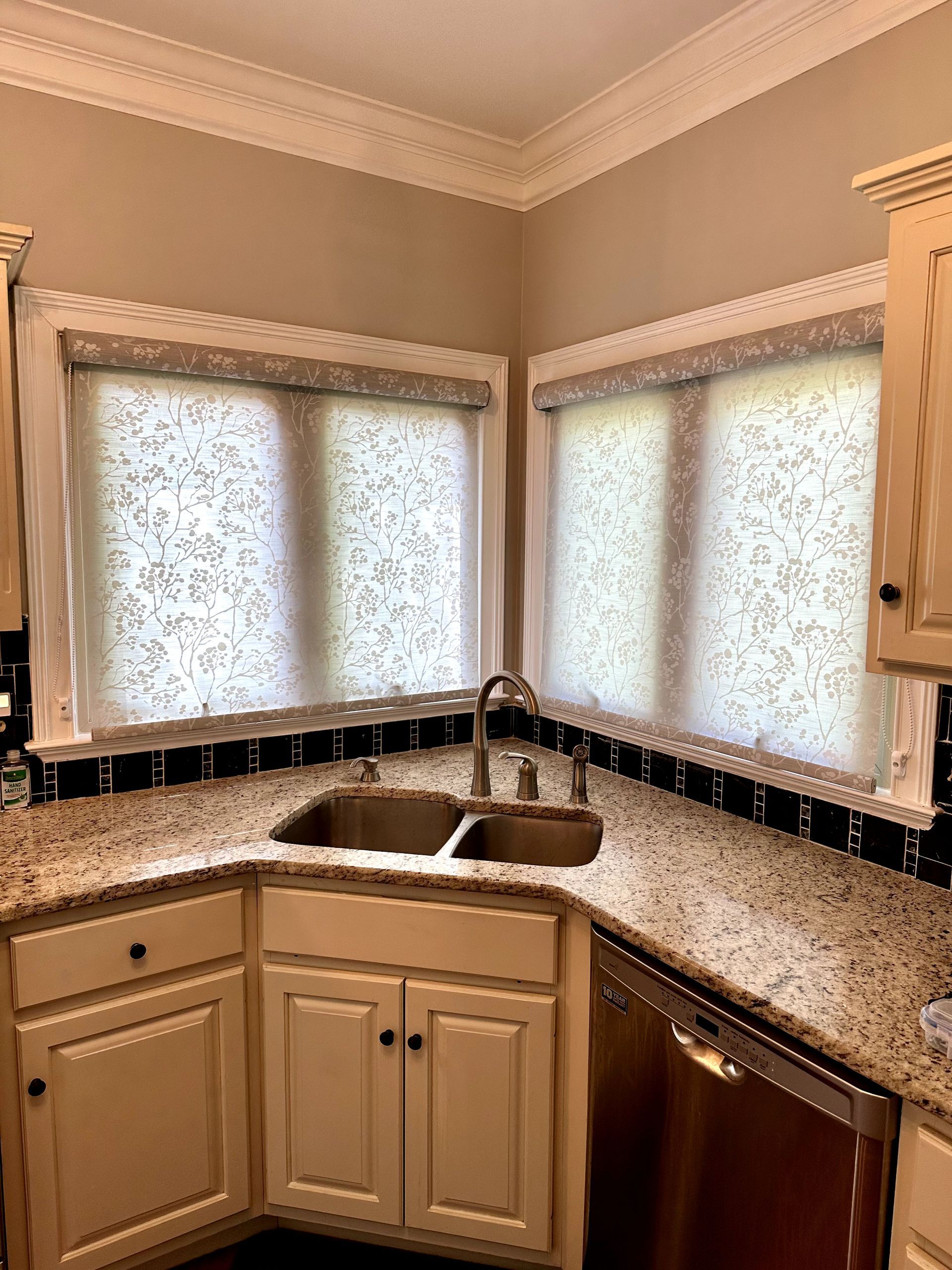 Kitchen corner with sink, granite countertops, cream cabinets, and patterned window shades.