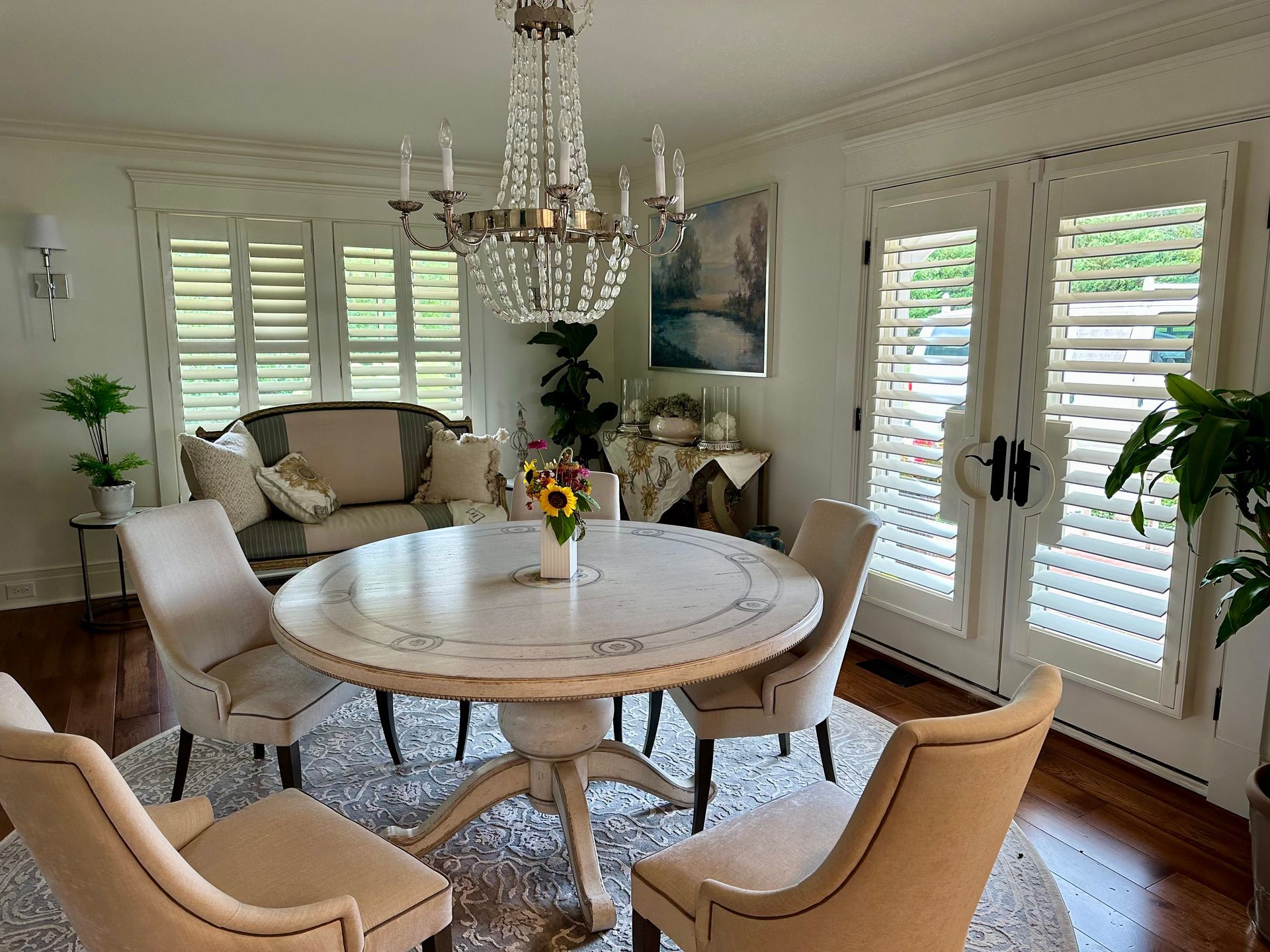 Elegant dining room with round table, four chairs, crystal chandelier, and shuttered windows.