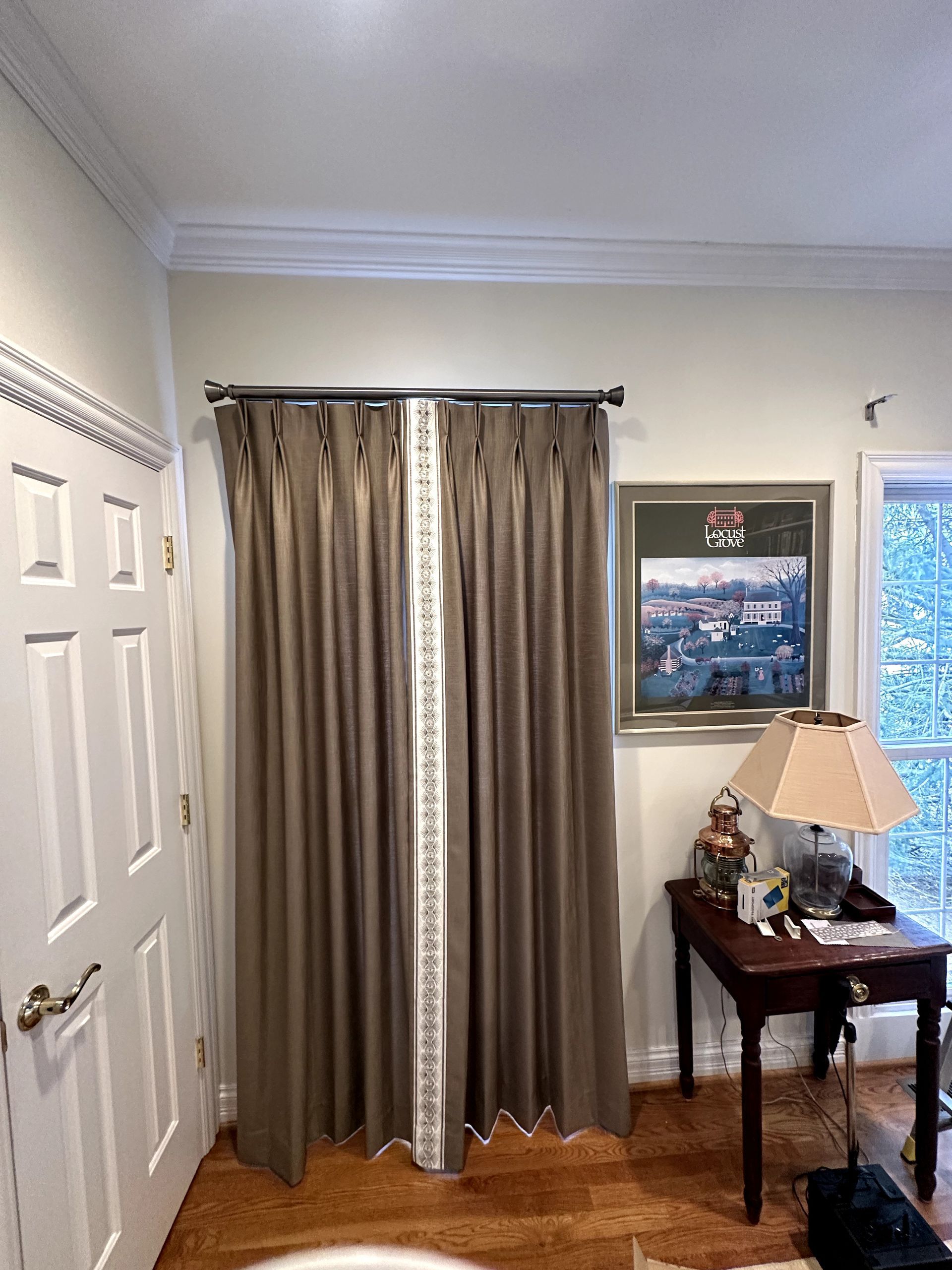 Brown curtains with white trim, hung on a rod. A painting, lamp, and table are visible. A door is on the left.