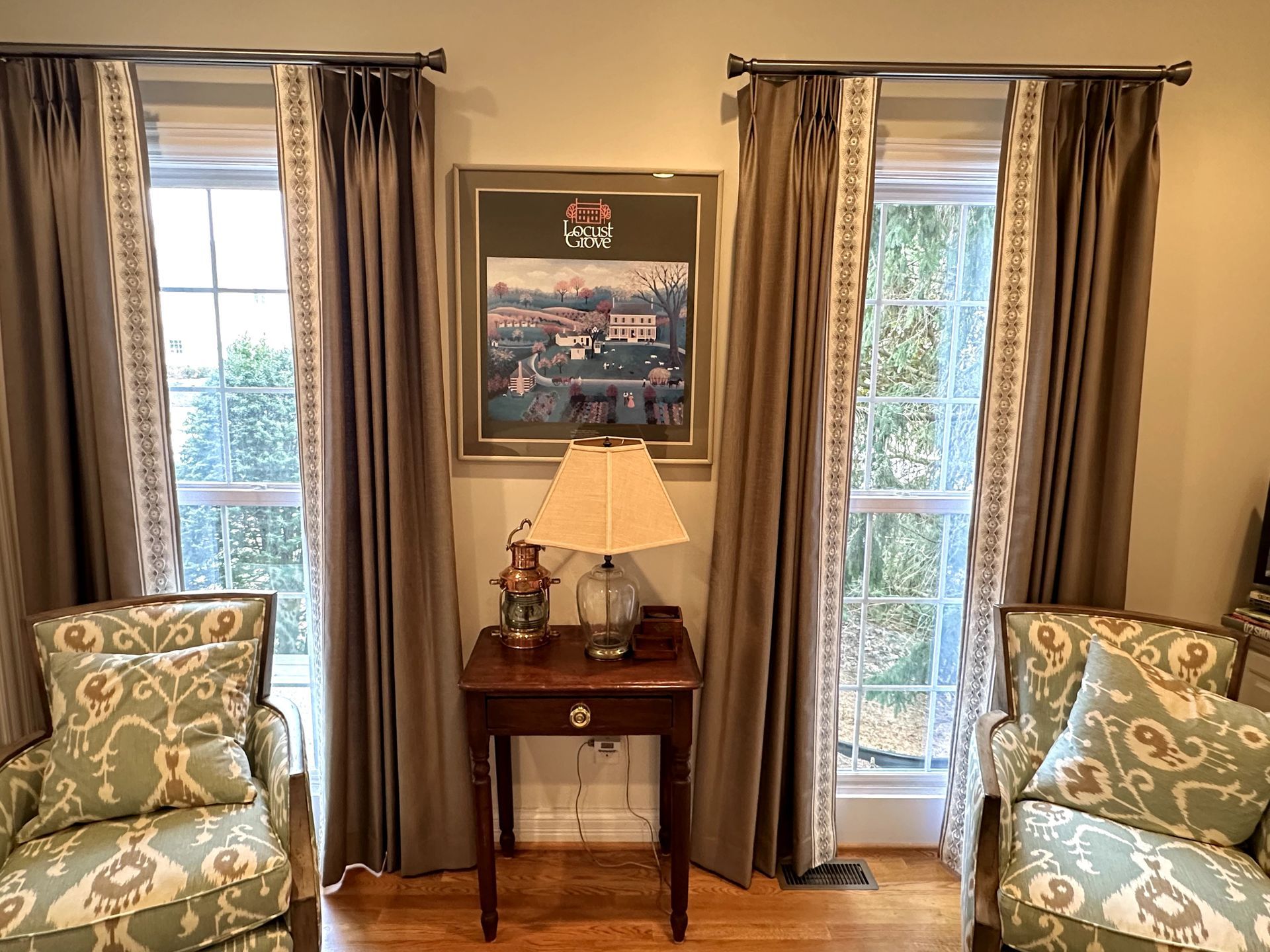 Two windows with brown curtains flanking a painting and table with lamp. Two patterned chairs are in front.