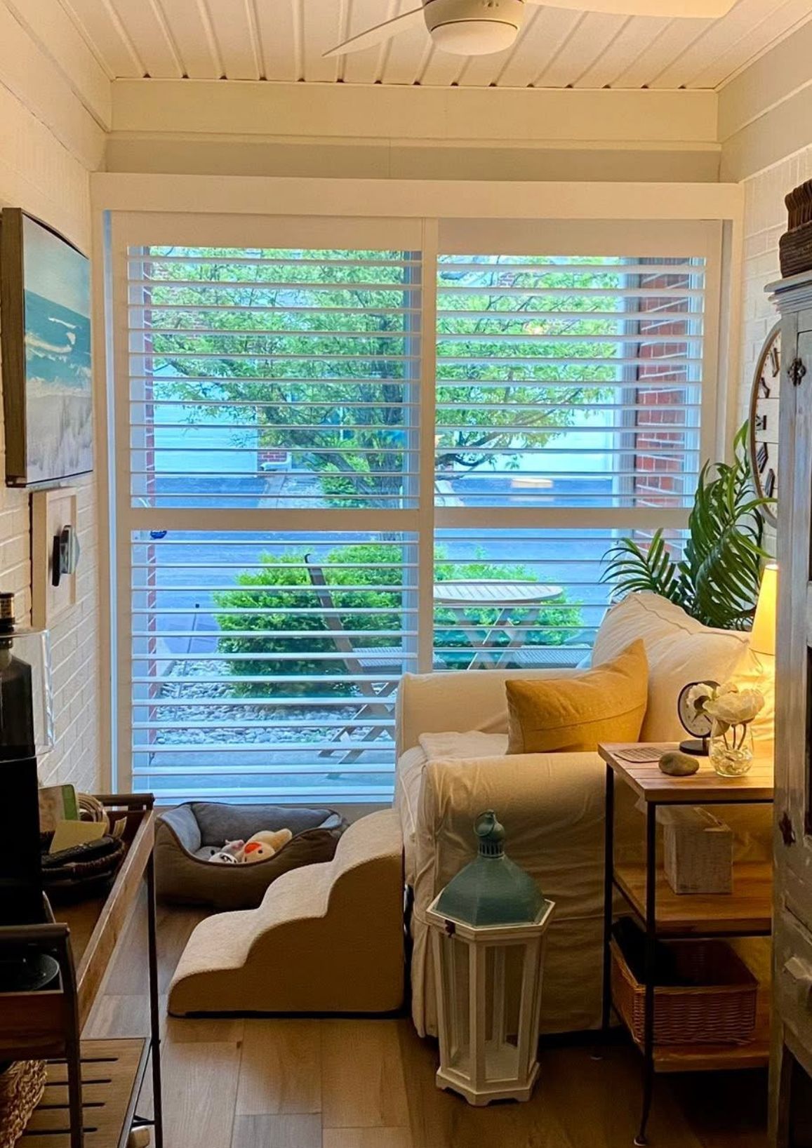 Cozy sunroom with white shutters, a sofa, and a dog bed. Bright natural light and a view of greenery.