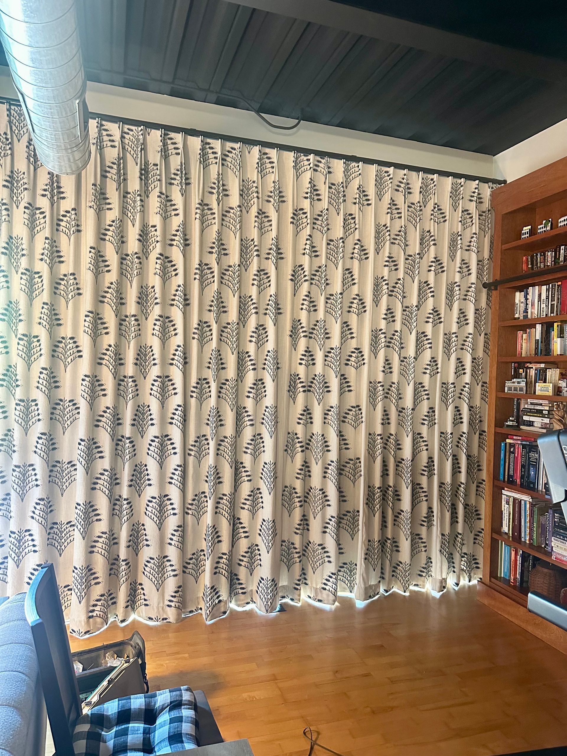 Beige curtains with black floral pattern against a dark ceiling, next to a bookcase on a hardwood floor.