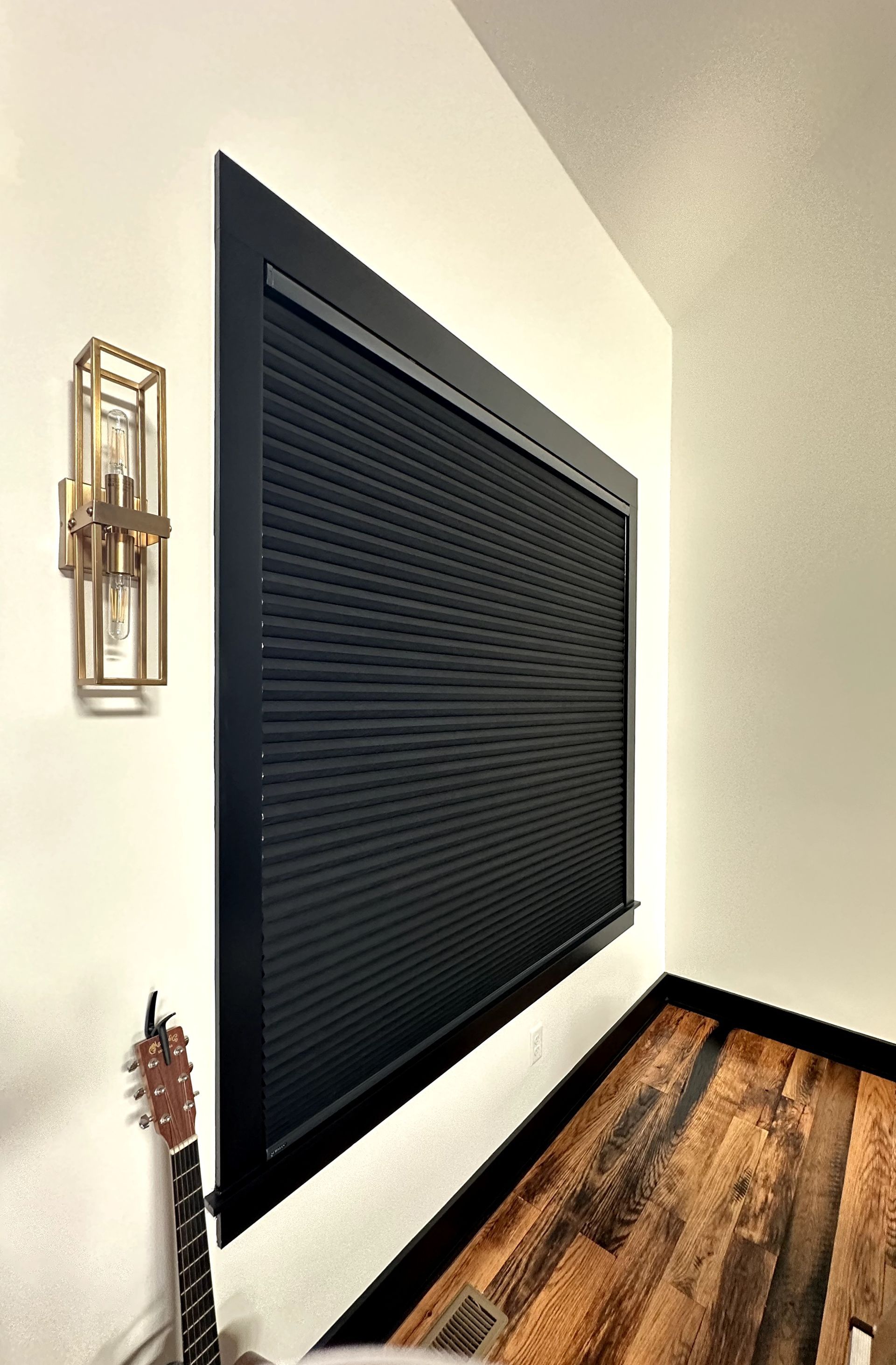 Black window with blinds framed in black trim, next to a gold sconce and guitar.