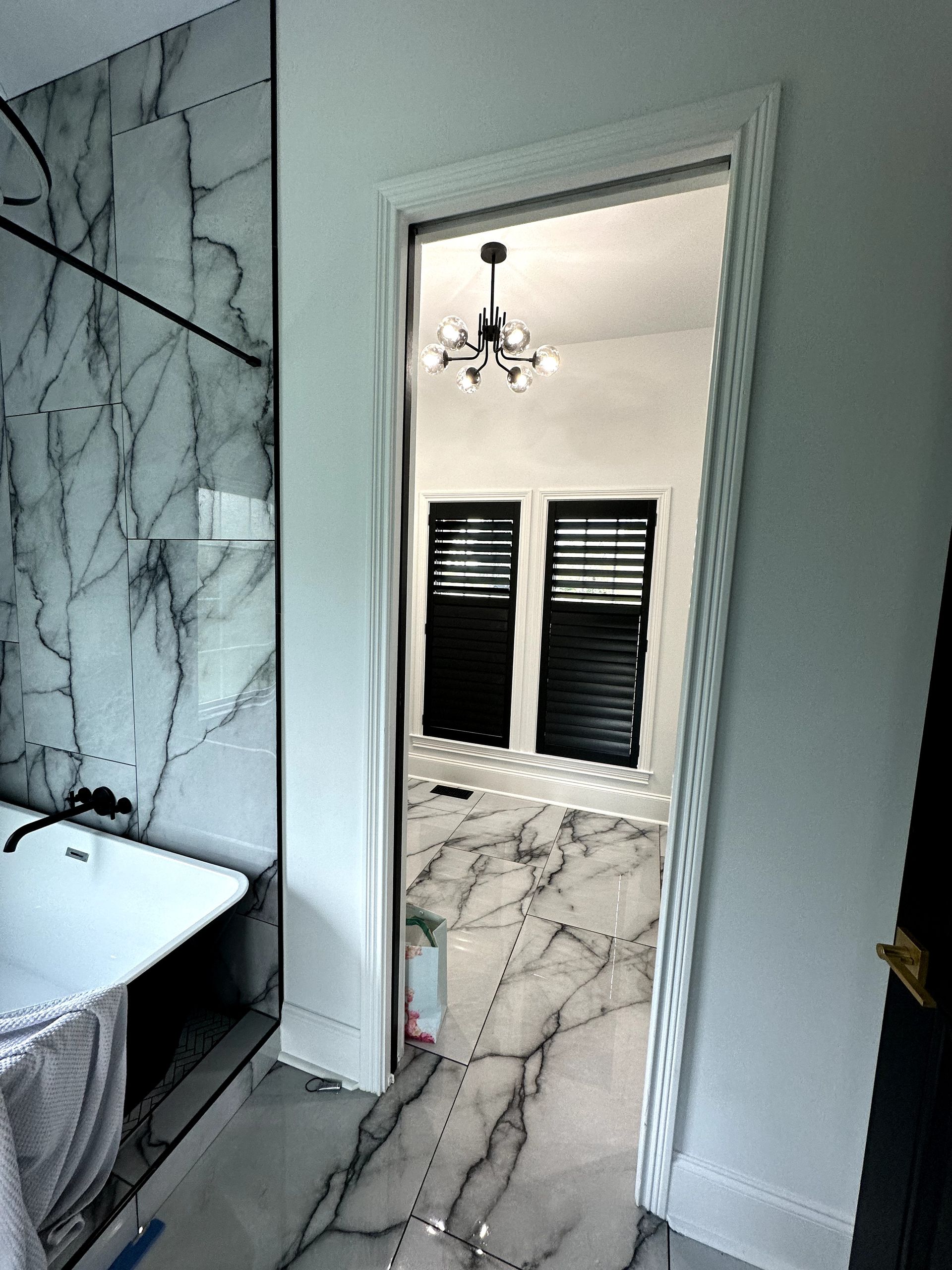 Bathroom with marble floors and walls, a view of another room with black shutters and a chandelier.