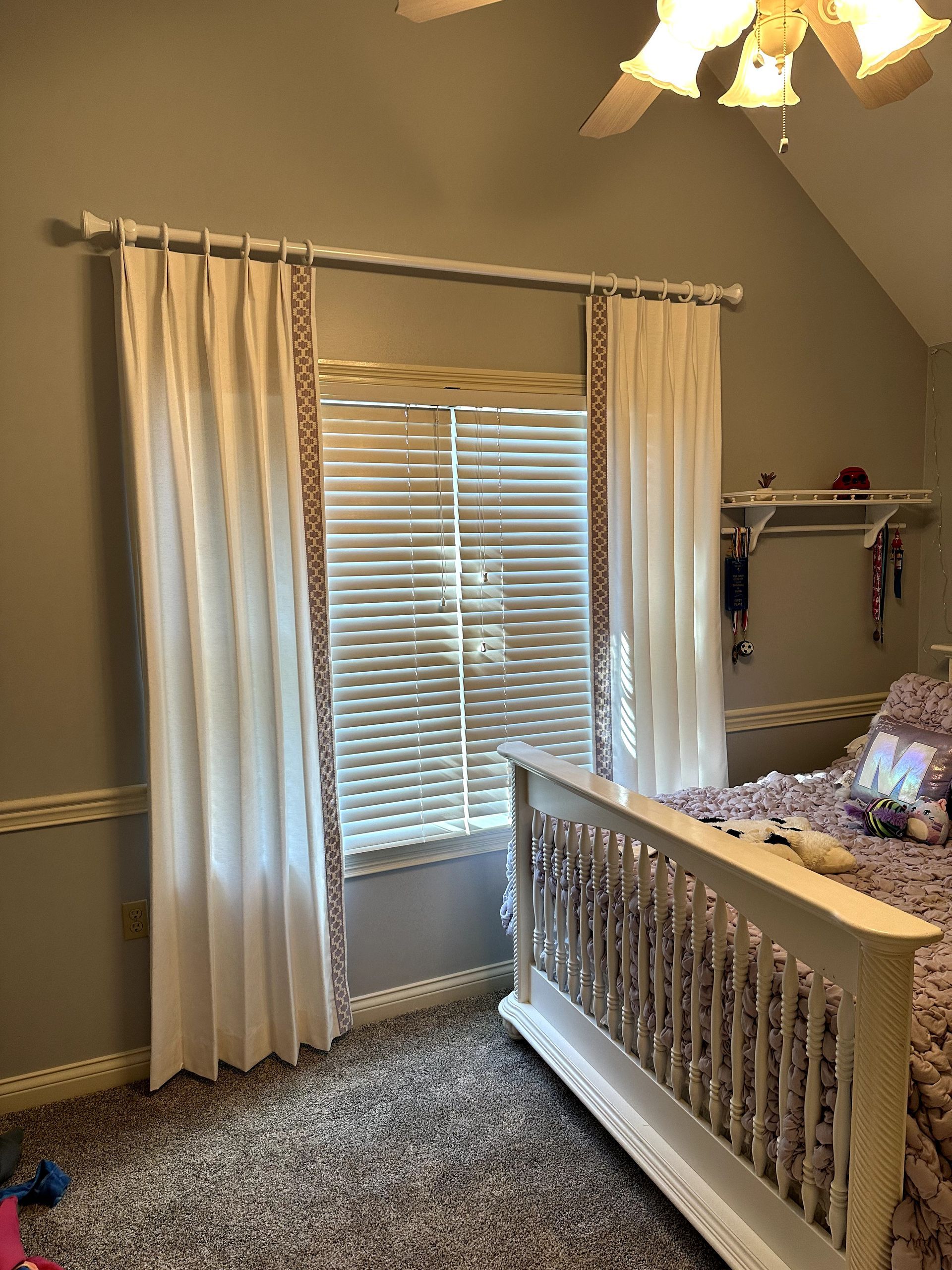 White curtains frame a window with blinds, next to a bed in a bedroom.