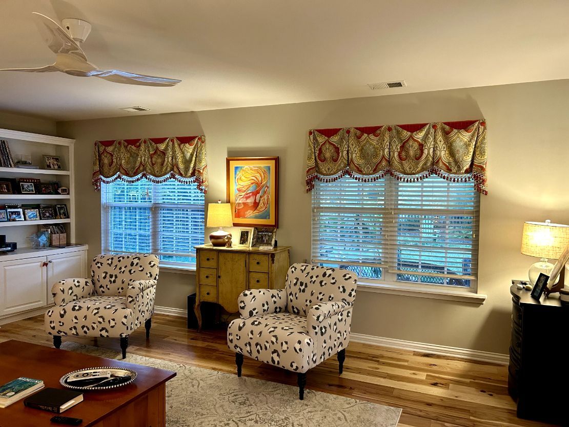 Living room with floral chairs, wooden cabinet, lamps, and sunlit windows with patterned valances