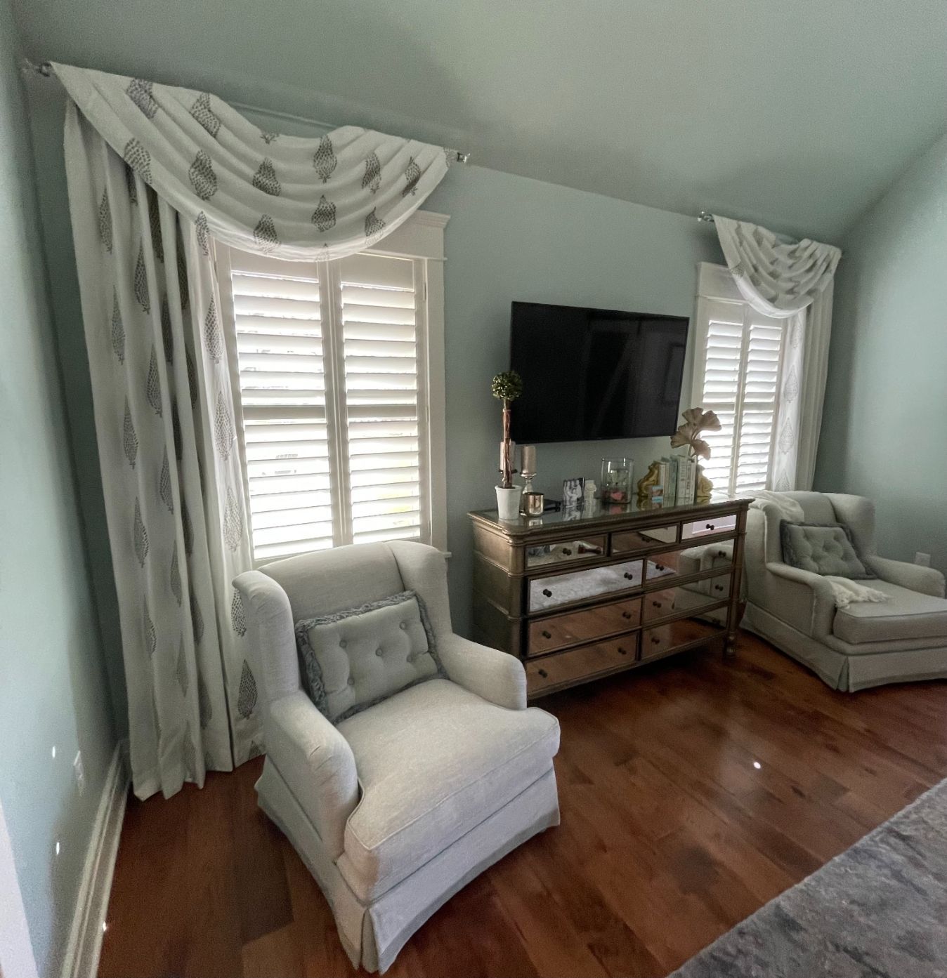 Bright living room with pale armchairs, white curtains, wooden dresser, wall-mounted TV, and hardwood floors