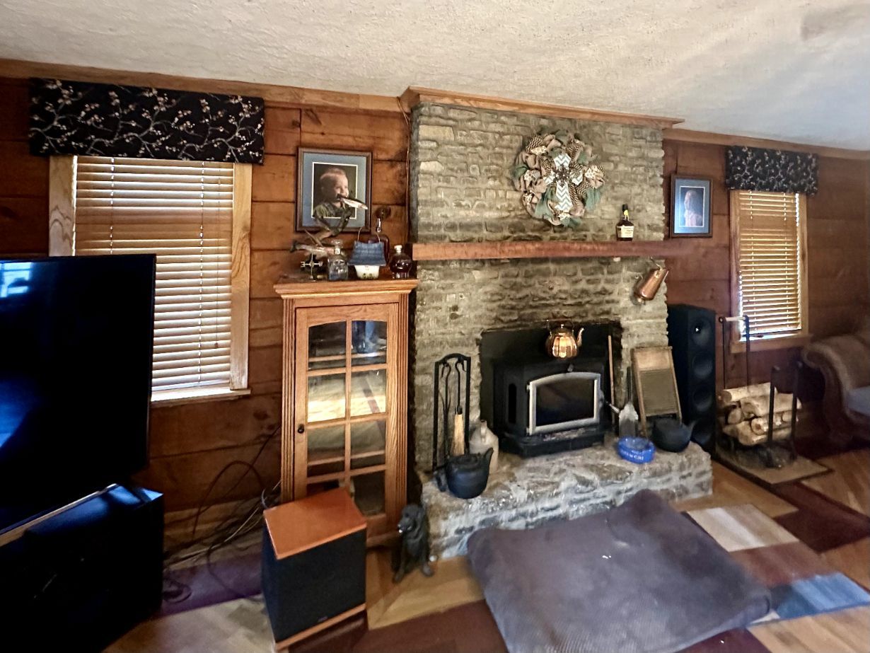 Cozy living room with stone fireplace, wood-paneled walls, TV, windows, and real wood blinds.