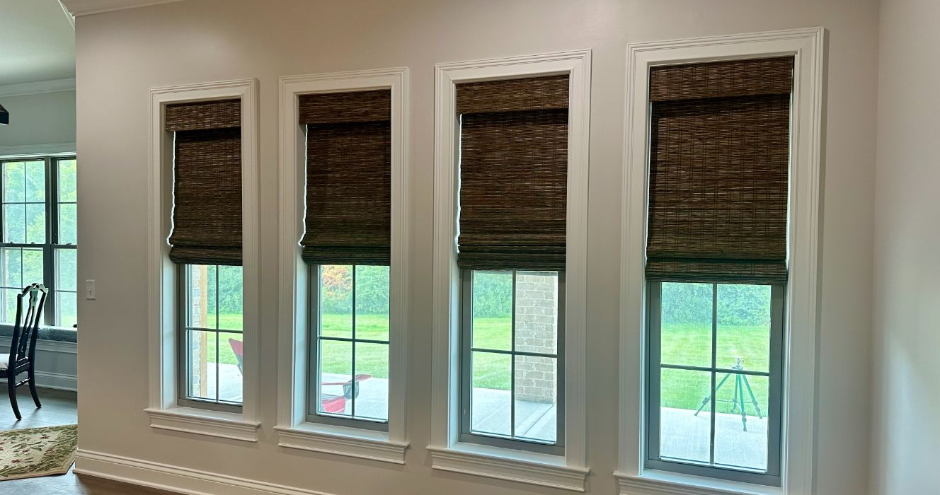 Four tall windows with woven shades in a bright room, overlooking a green lawn