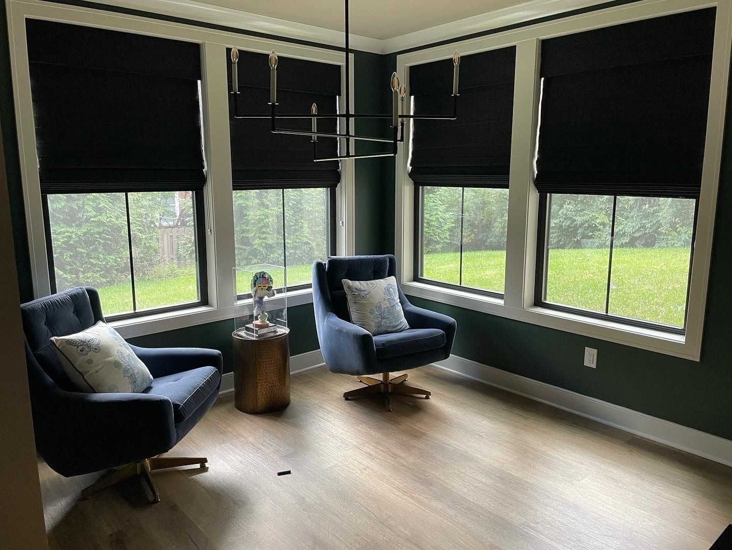 Cozy corner room with two blue chairs, a small table, large windows, and green lawn outside.