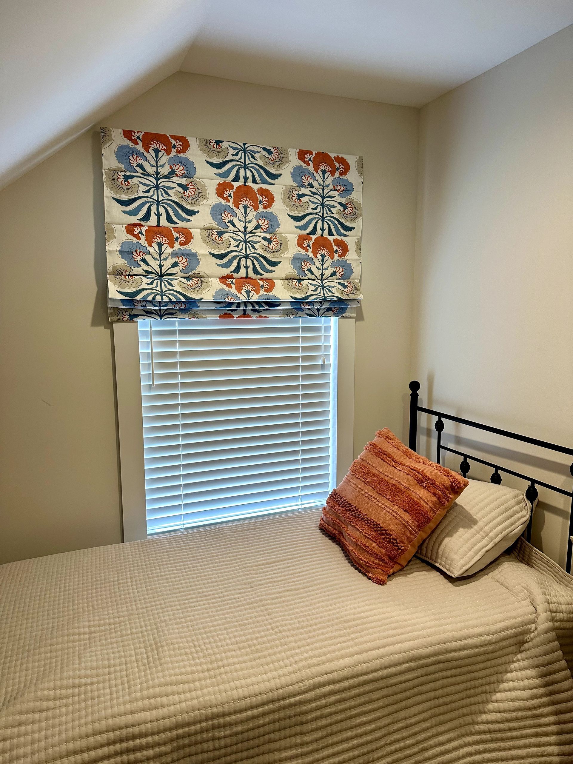 Bedroom with a twin bed, patterned Roman shade, and white blinds. Two pillows and a white coverlet on the bed.