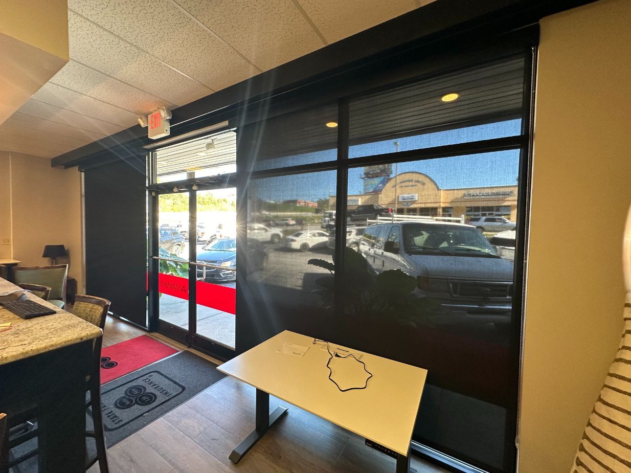 Interior view: Black roller shades cover large windows overlooking a parking lot. A table sits near the window.