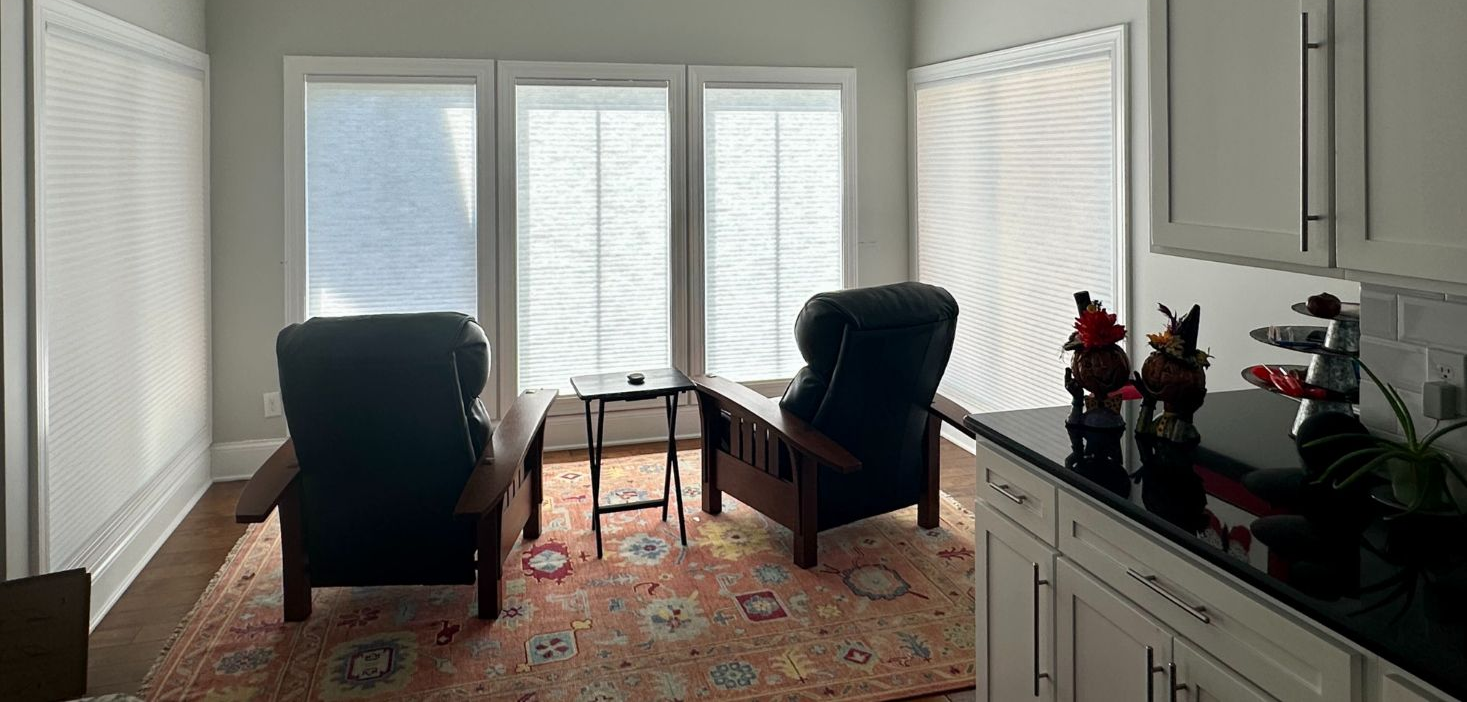 Two dark armchairs face a windowed corner in a sunlit room with light grey walls, a patterned rug, and kitchen cabinets.