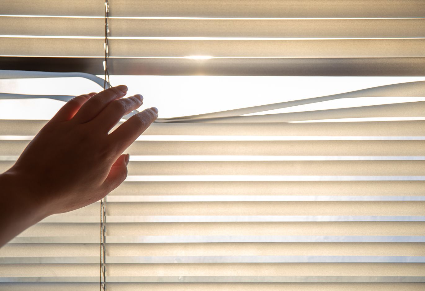 Hand parting window blinds, letting sunlight stream through.