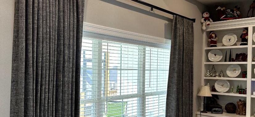 Window with gray curtains and white blinds beside a wall shelf of plates and decor.