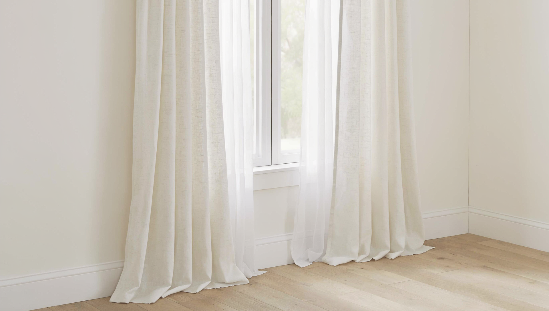 Light custom drapery framing a sunlit window in a minimalist room with pale walls and wood flooring