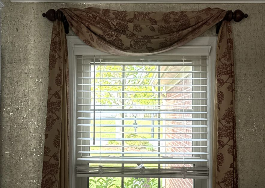 A window with white horizontal blinds, topped with a draped, patterned fabric valance and side panels on a dark rod.