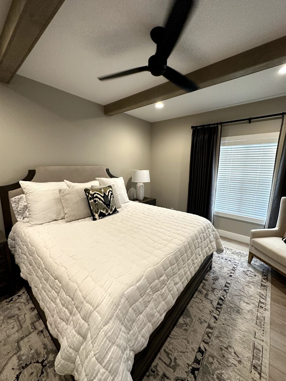Bright bedroom with a white bed, dark ceiling fan, lamp, and window with blinds and curtains.